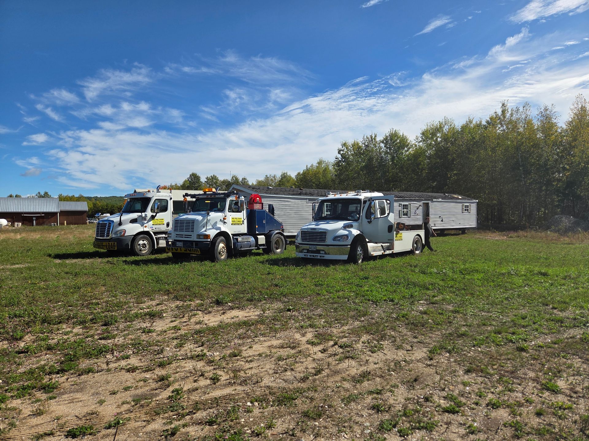 Three white industrial trucks are parked in a grassy field on a sunny day with a blue sky and a line of trees in the back.