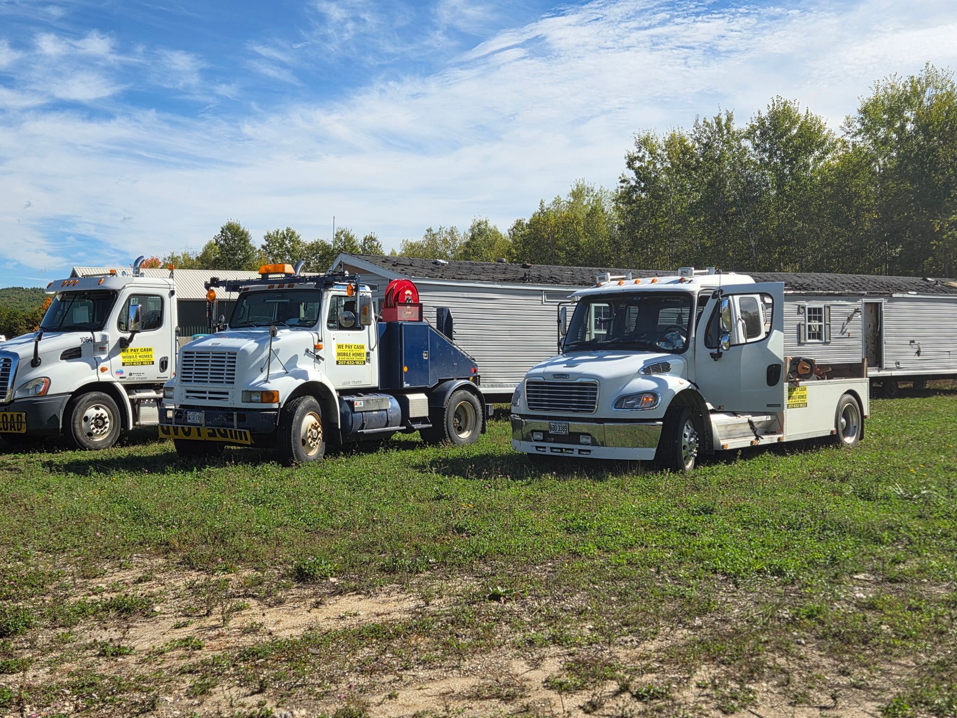 Three white tow trucks are parked in a grassy field in front of a white trailer on a bright, sunny day.