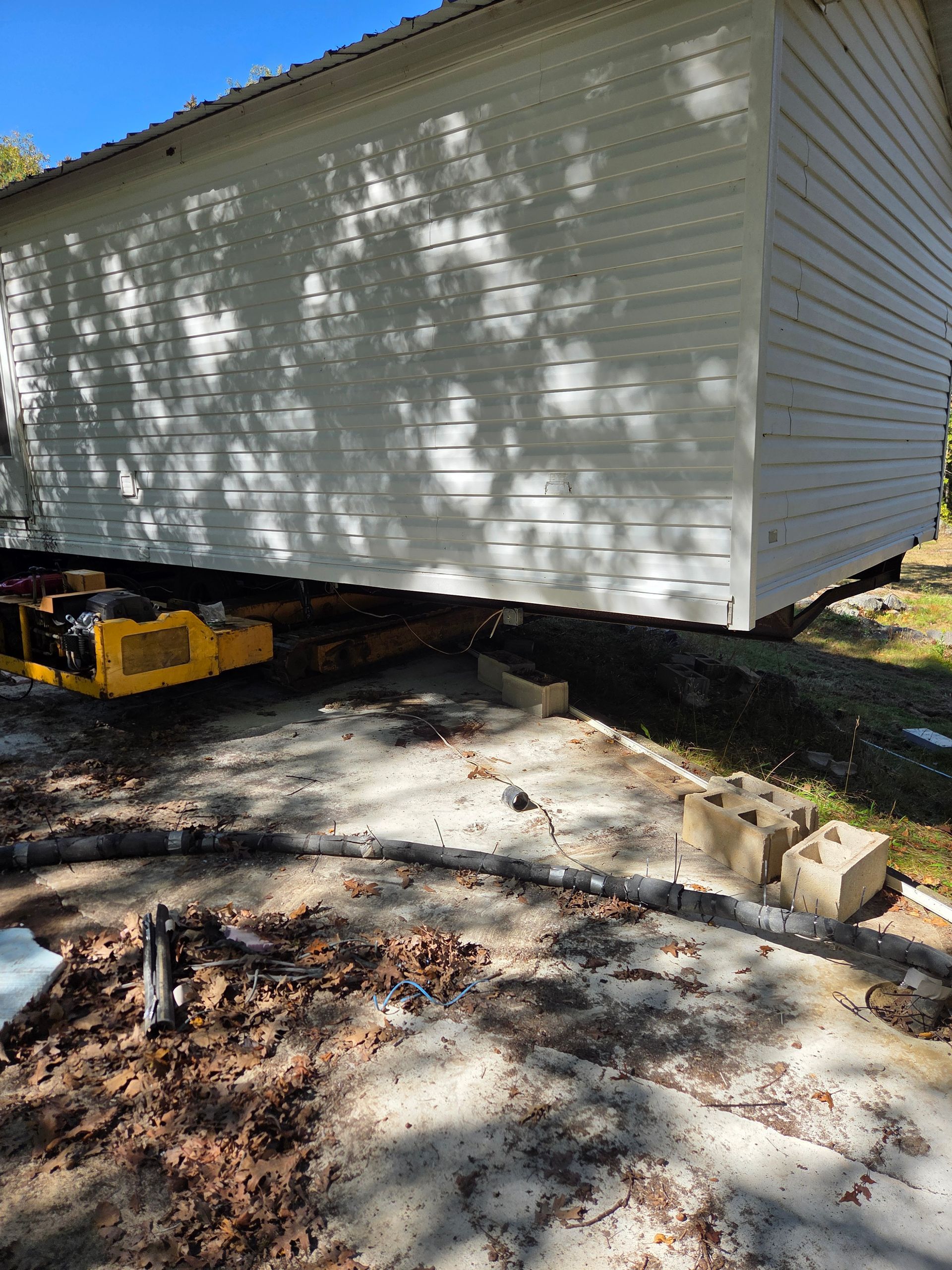 A white building sits on a yellow hydraulic house-moving trailer on a dirt ground covered with leaves.