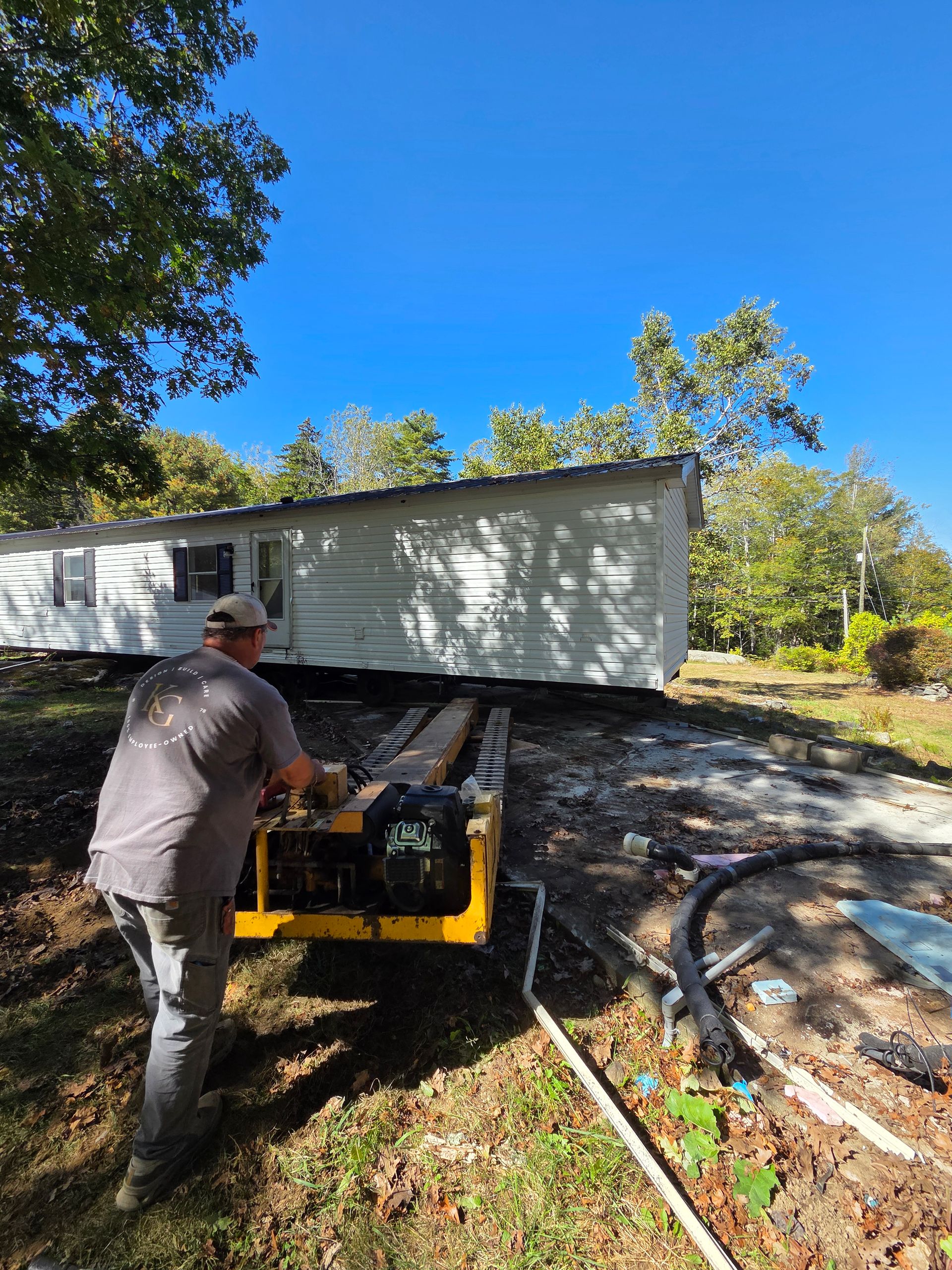 A person operates a yellow motorized dolly to move a white manufactured home across a grassy, wooded lot.