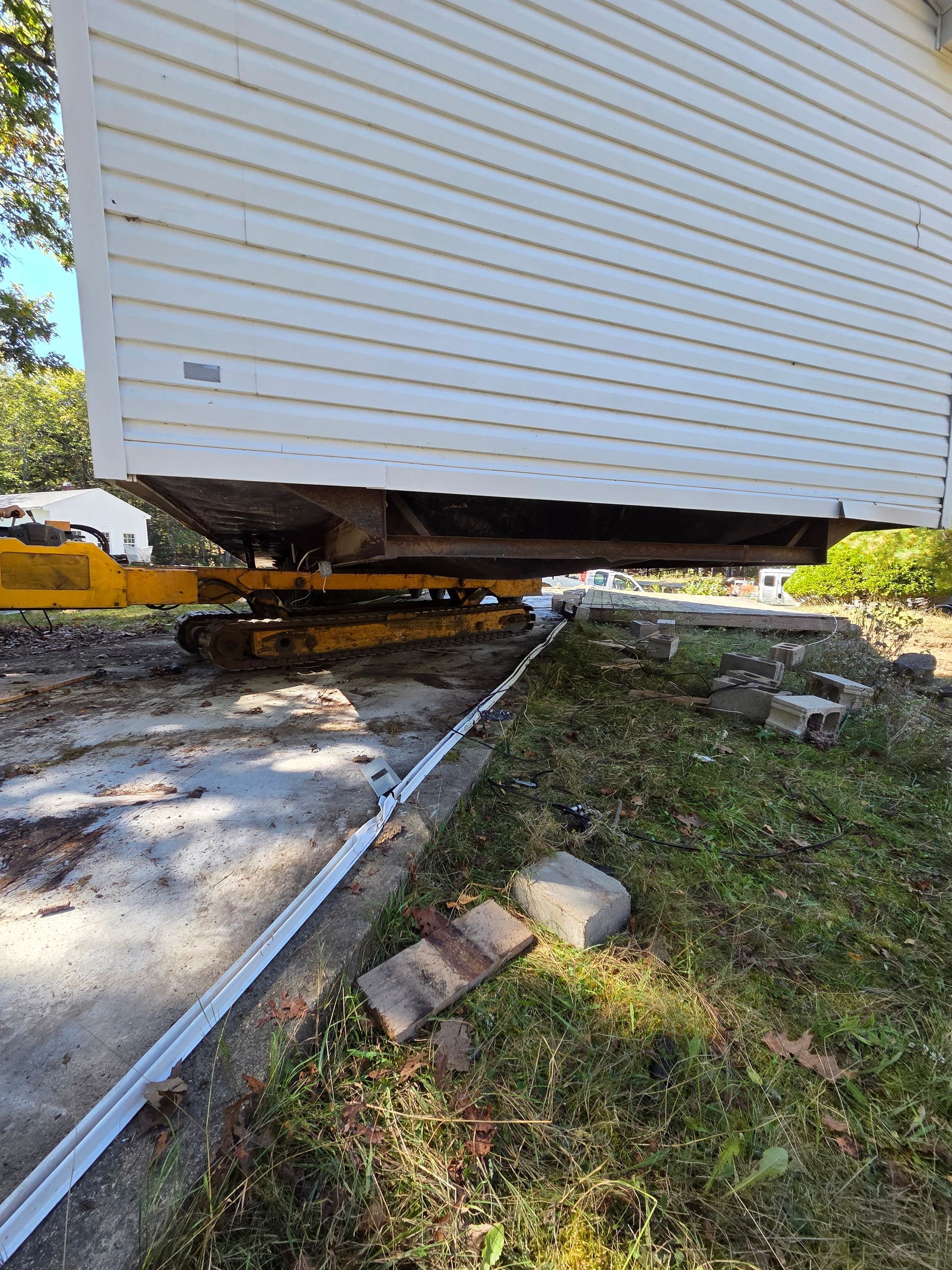 A large white house structure on a yellow metal transport trailer being moved along a concrete driveway outdoors.