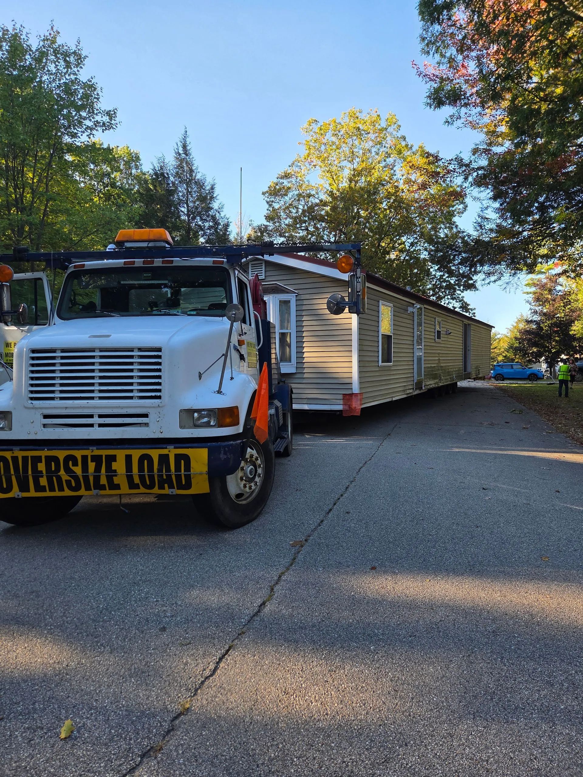 Tow truck hauling a white modular home trailer on a tree-lined residential street