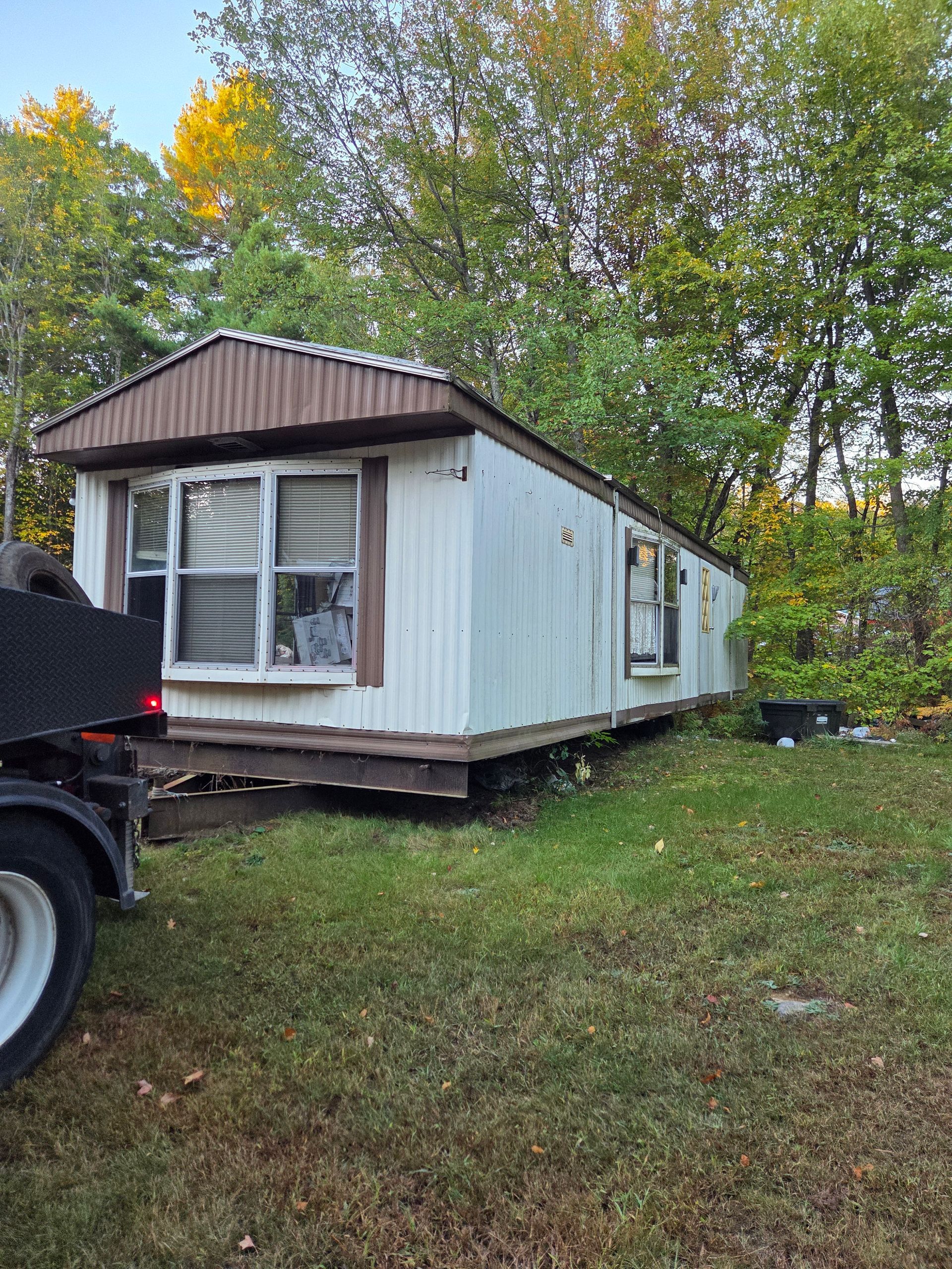 A weathered, single-wide mobile home parked on grass, partially hitched to a truck in a wooded area.