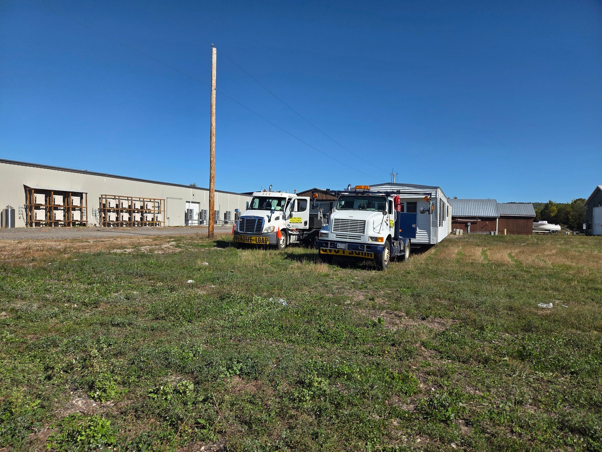 Two white commercial trucks parked on a grass field, one towing a mobile building next to a large warehouse.