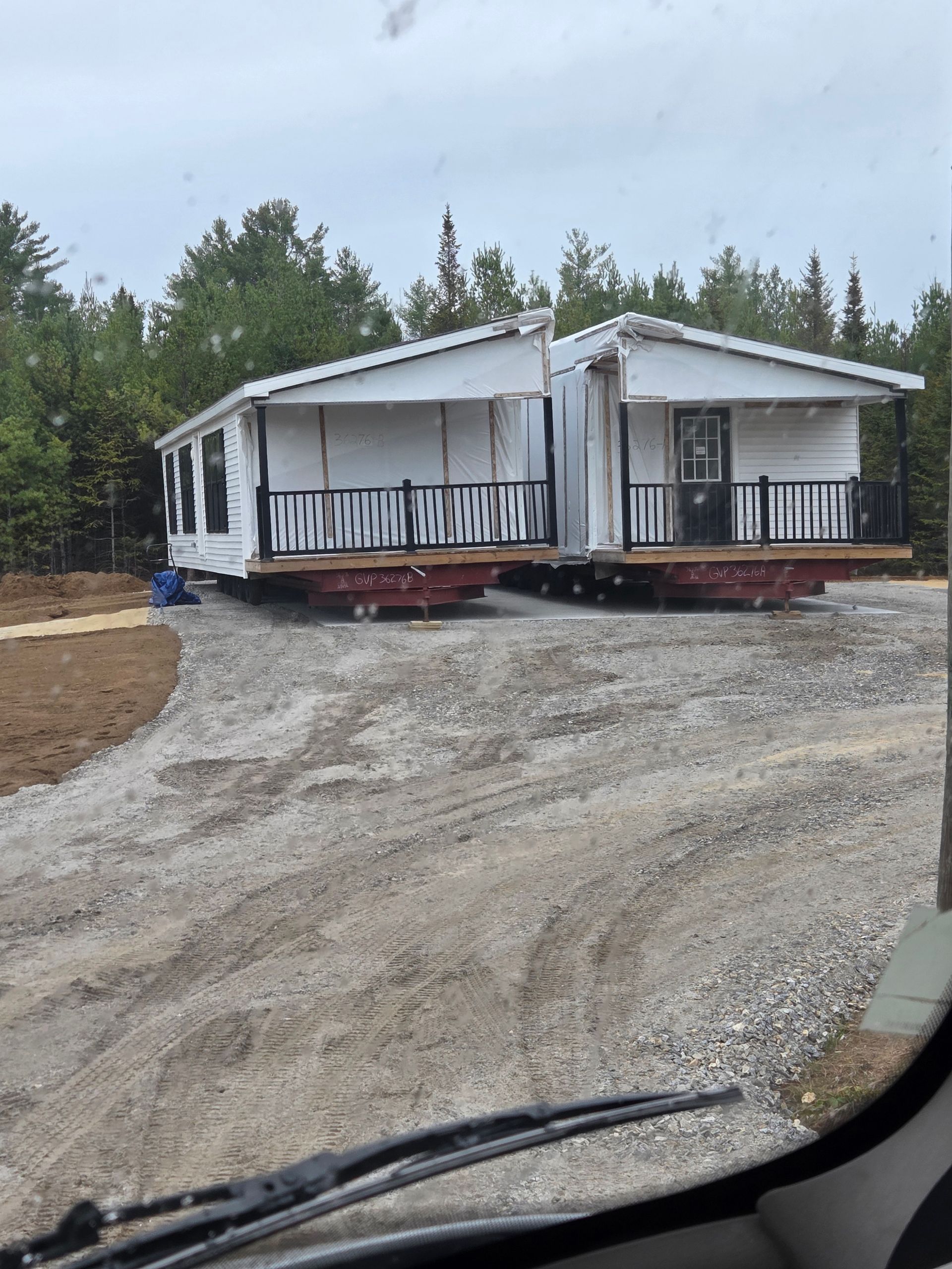 Two modular homes on trailers sit side-by-side on a gravel lot in a wooded area, awaiting installation.