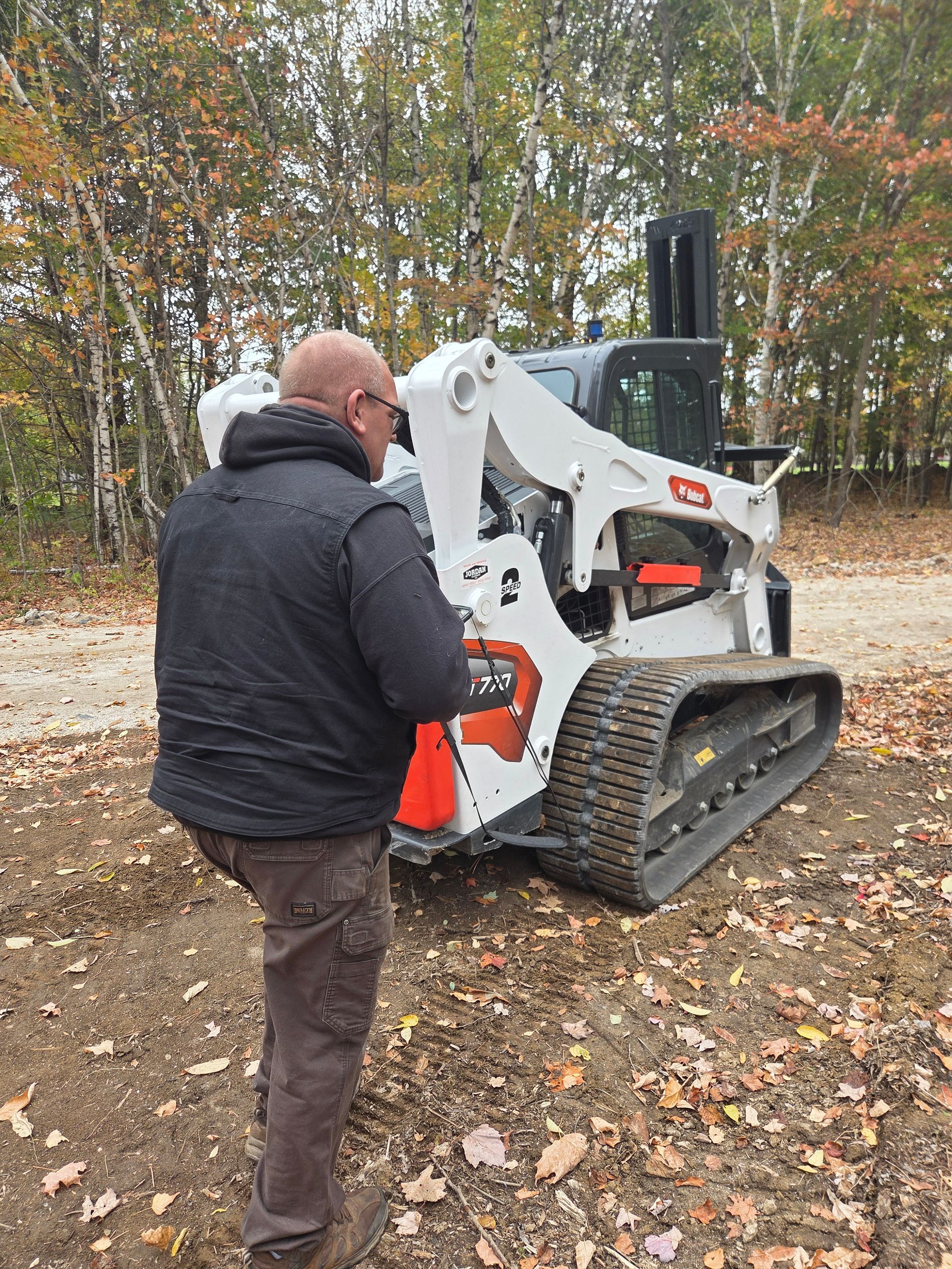 A person in a black jacket and brown pants stands next to a white Bobcat compact track loader in a wooded, leafy area.