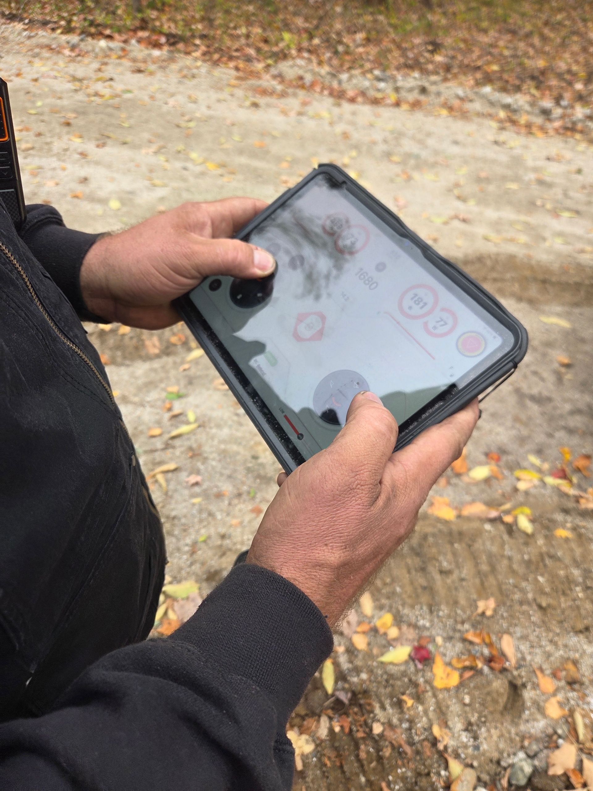 A pair of hands holds a tablet displaying a control interface outdoors against a backdrop of dirt and fallen leaves.