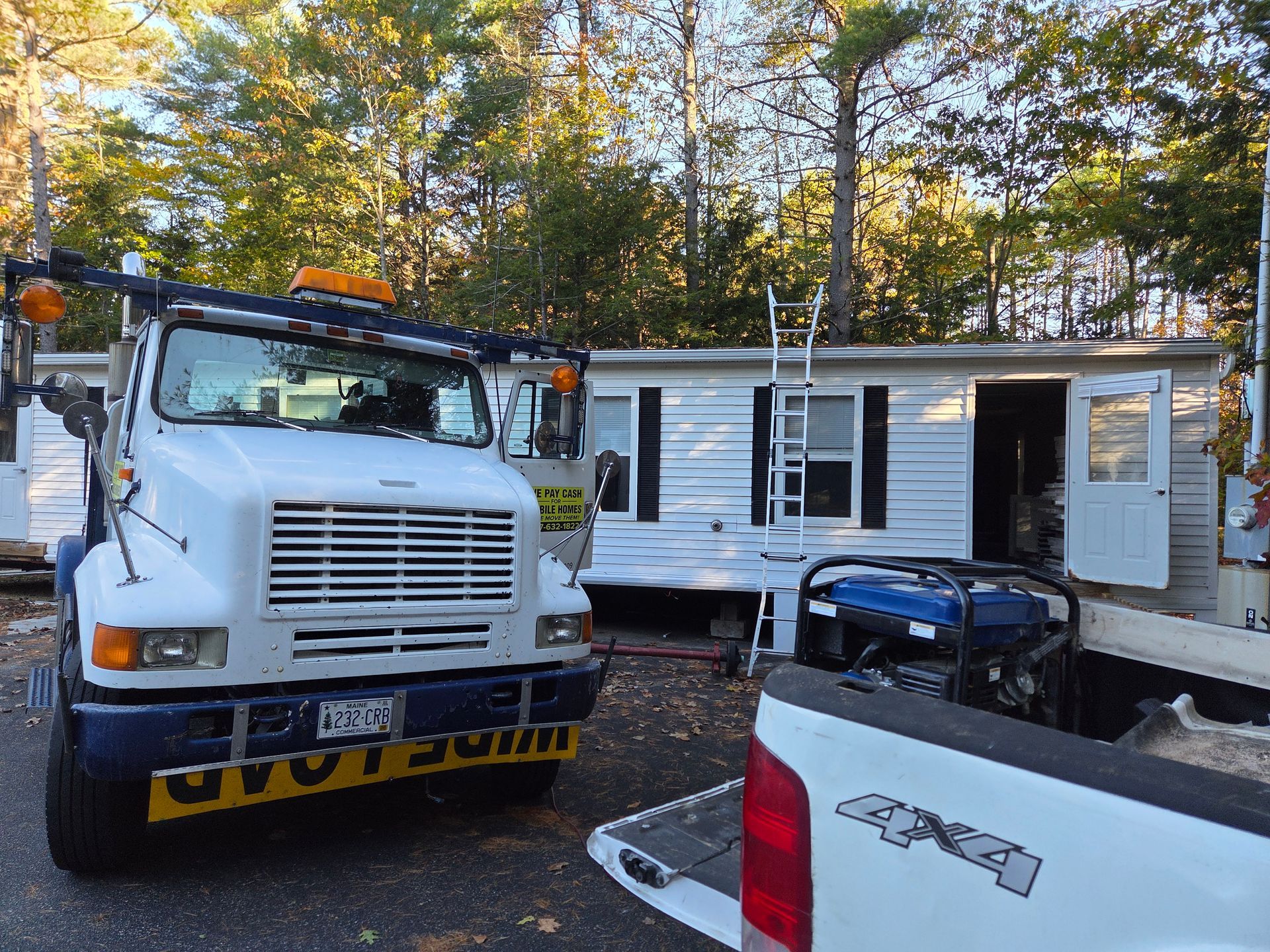 A white tow truck is parked in front of a mobile home with an open door and a ladder leaning against its side.