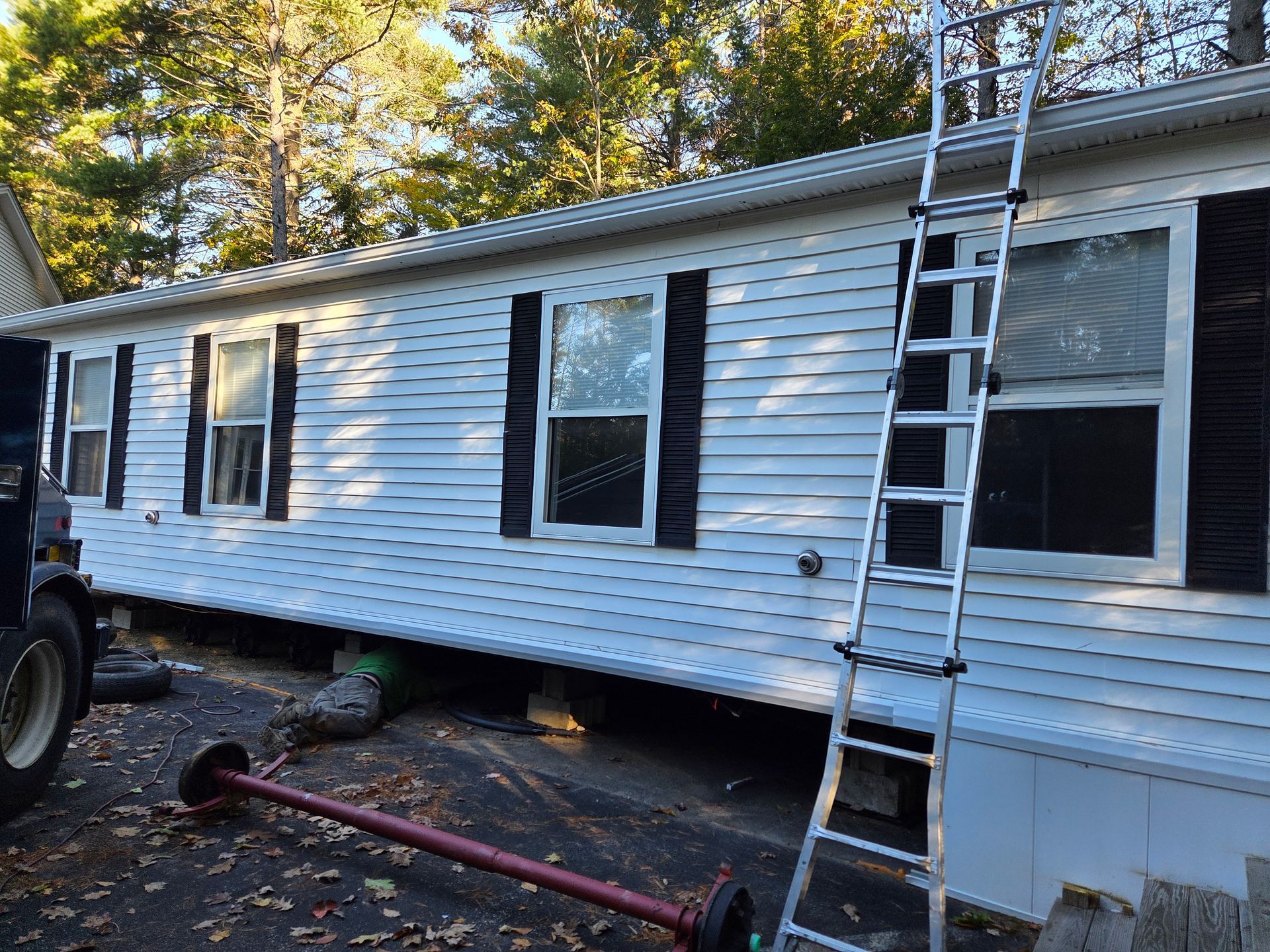A white mobile home with black shutters, an exterior ladder against the side, and a detached metal axle on the ground.