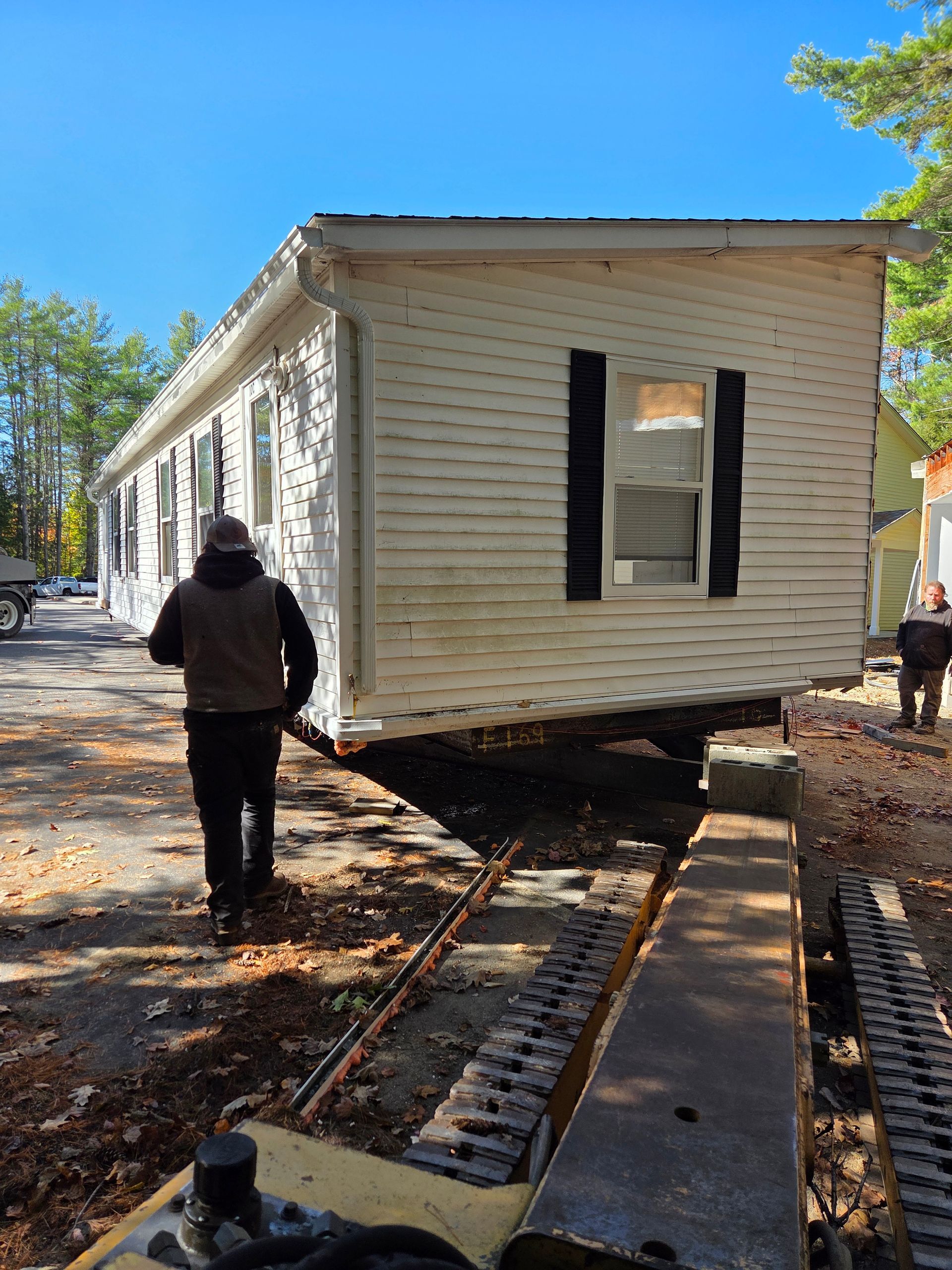A mobile home being relocated by a heavy machine on a site with trees under a blue sky.