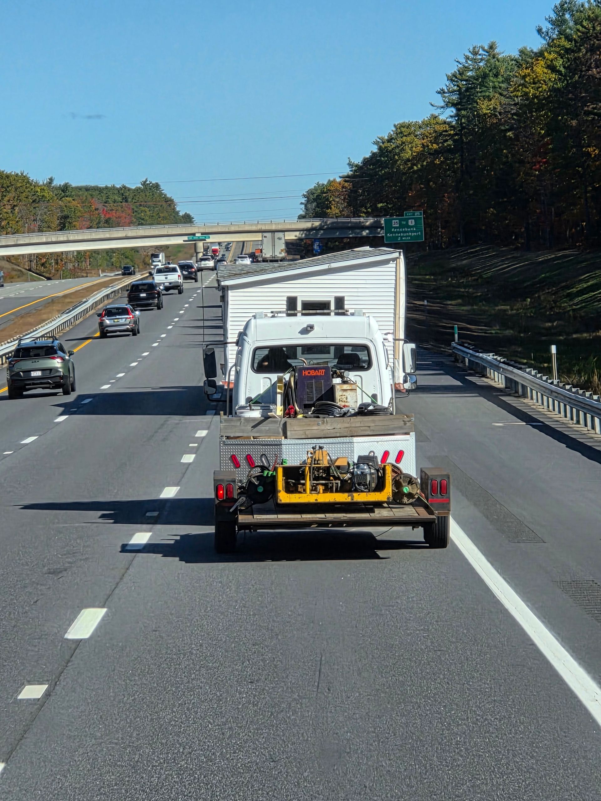 A view from behind a white truck carrying a tall, rectangular shed or structure on a highway with other cars nearby.