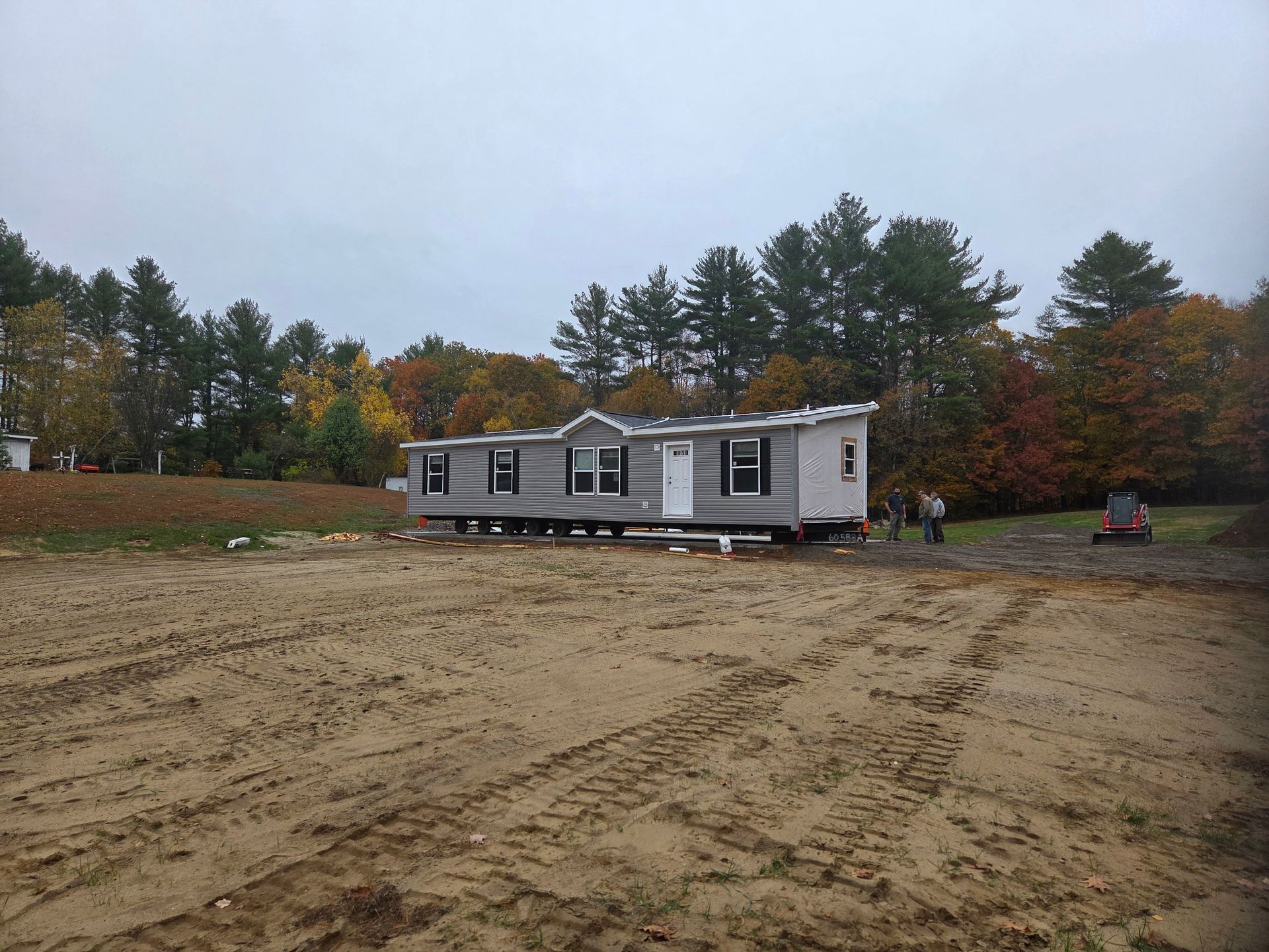 A grey manufactured home sits on a prepared sandy lot in front of a tree-lined backdrop under a cloudy sky.