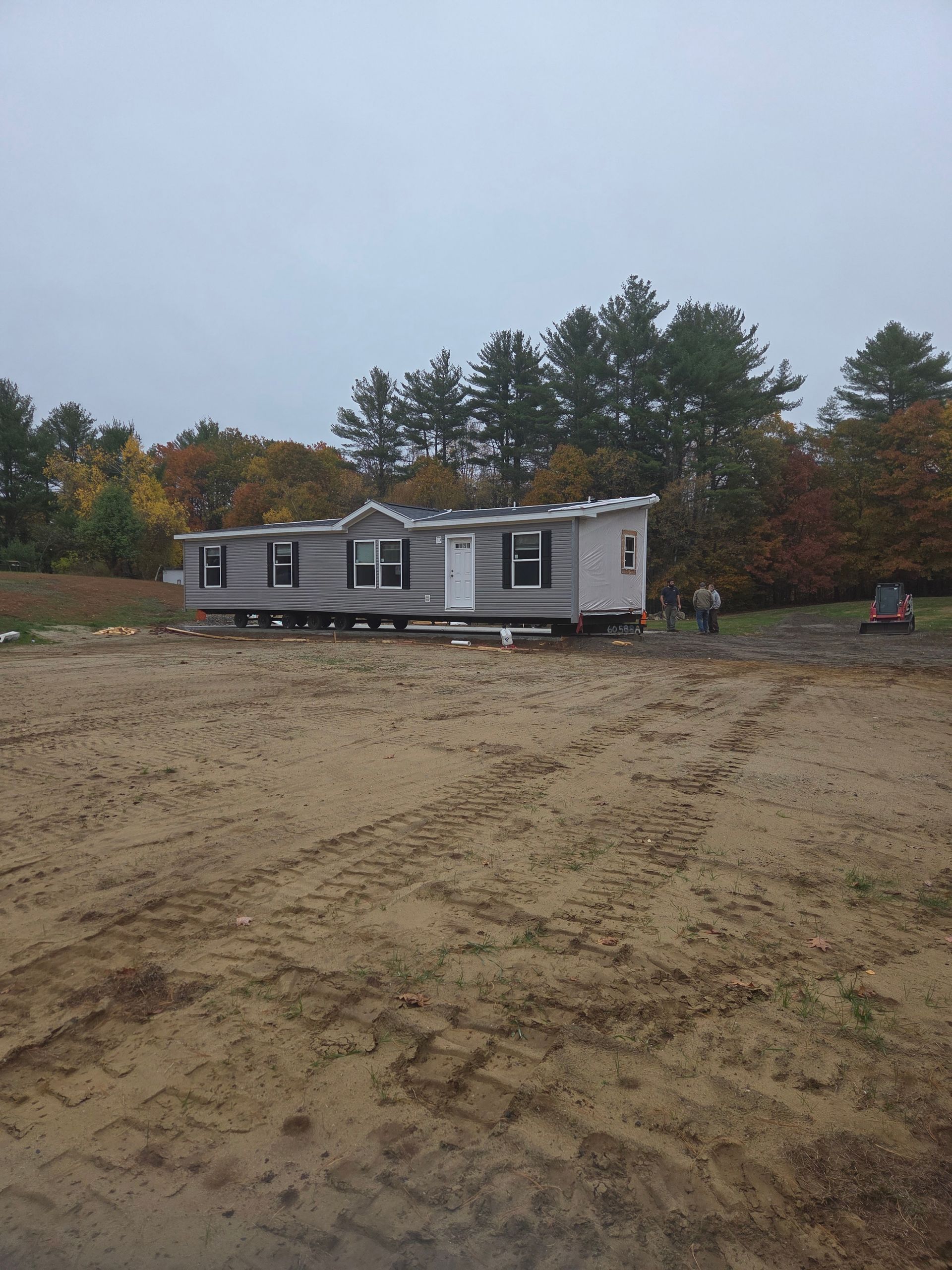 A grey manufactured home sits on a dirt lot in front of a tree line during an overcast day.