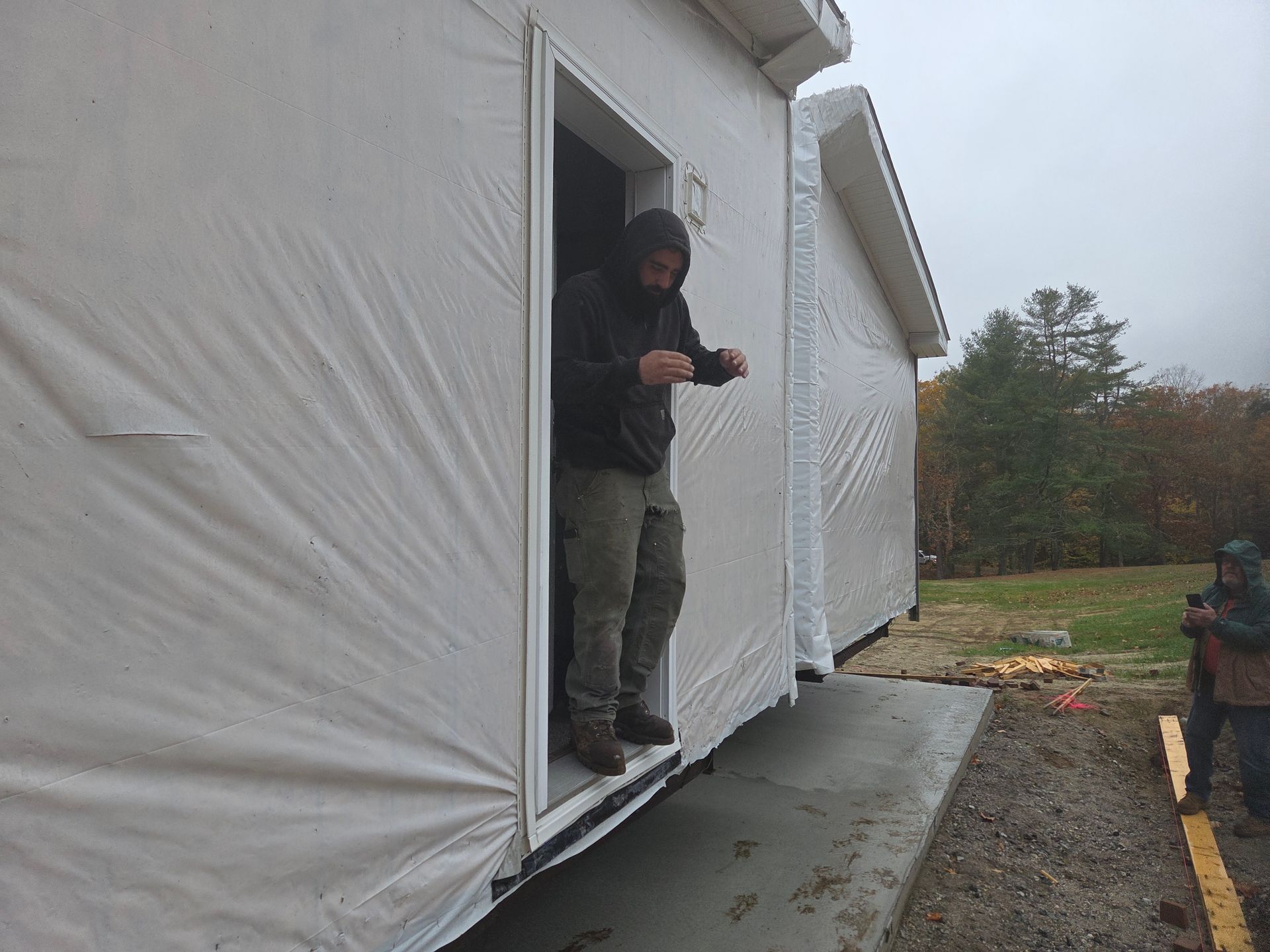 A person in a hooded sweatshirt exits a modular home wrapped in white protective sheeting onto a concrete pad.
