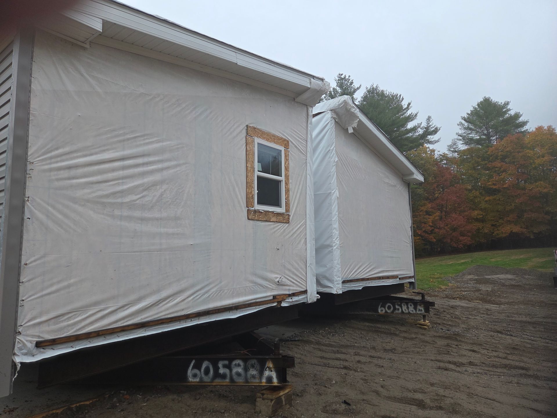 Two modular home sections on metal transport trailers, covered in protective white plastic wrap, sit on a dirt lot.