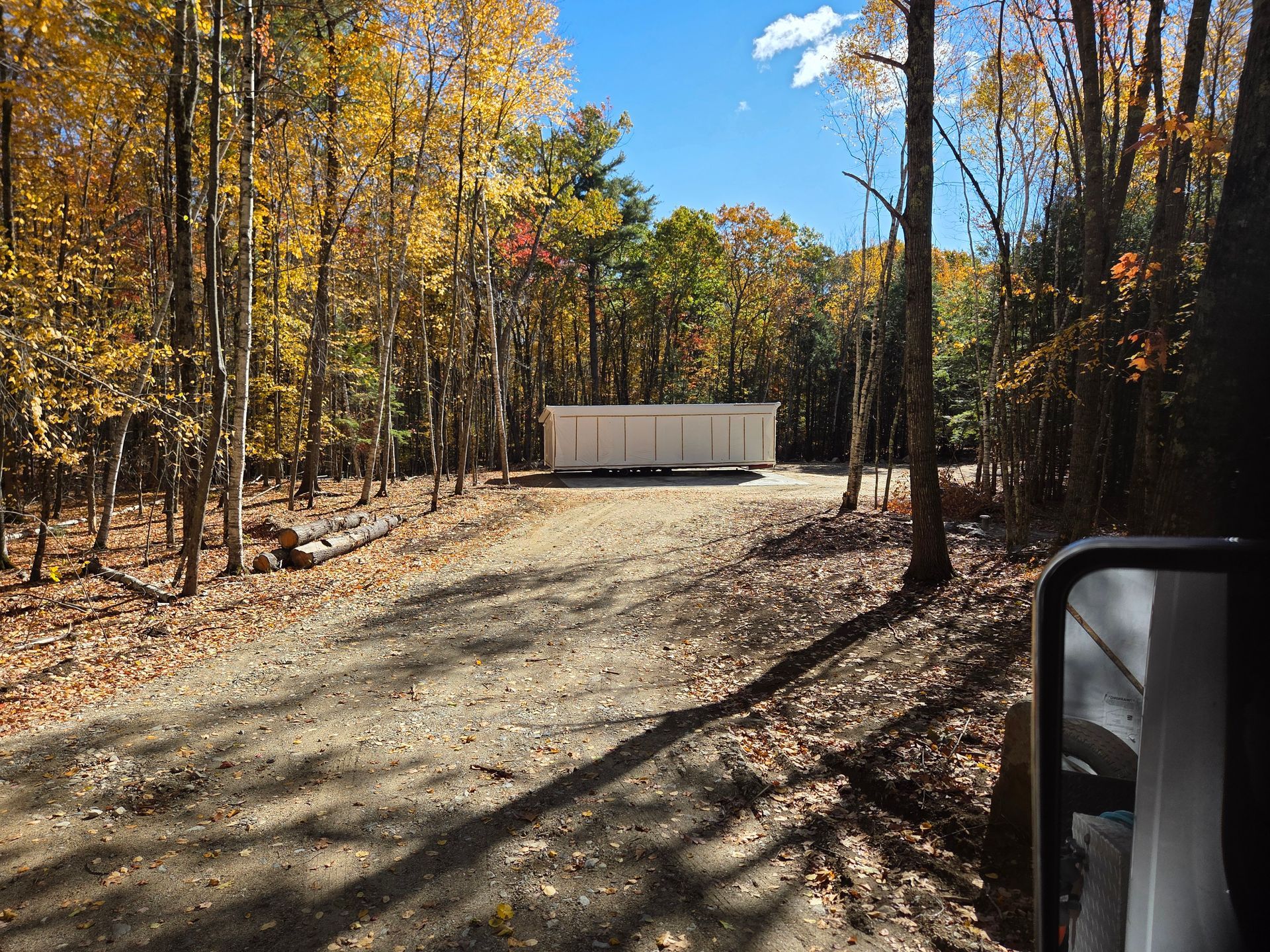 A white rectangular storage unit sits at the end of a gravel driveway in a wooded area with autumn foliage.
