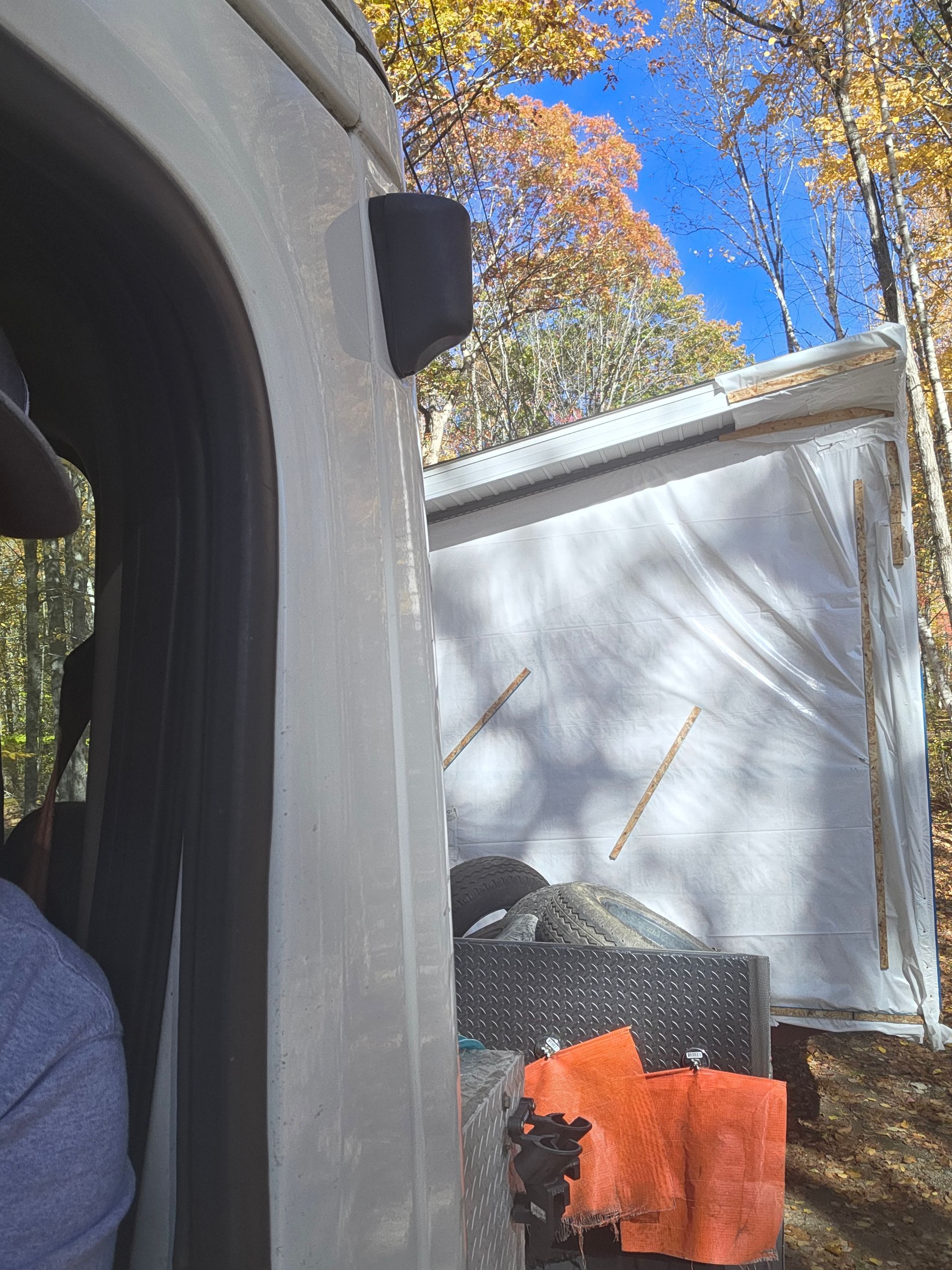 A view from inside a vehicle looking out at a white structure and orange gear in a wooded area with autumn trees.