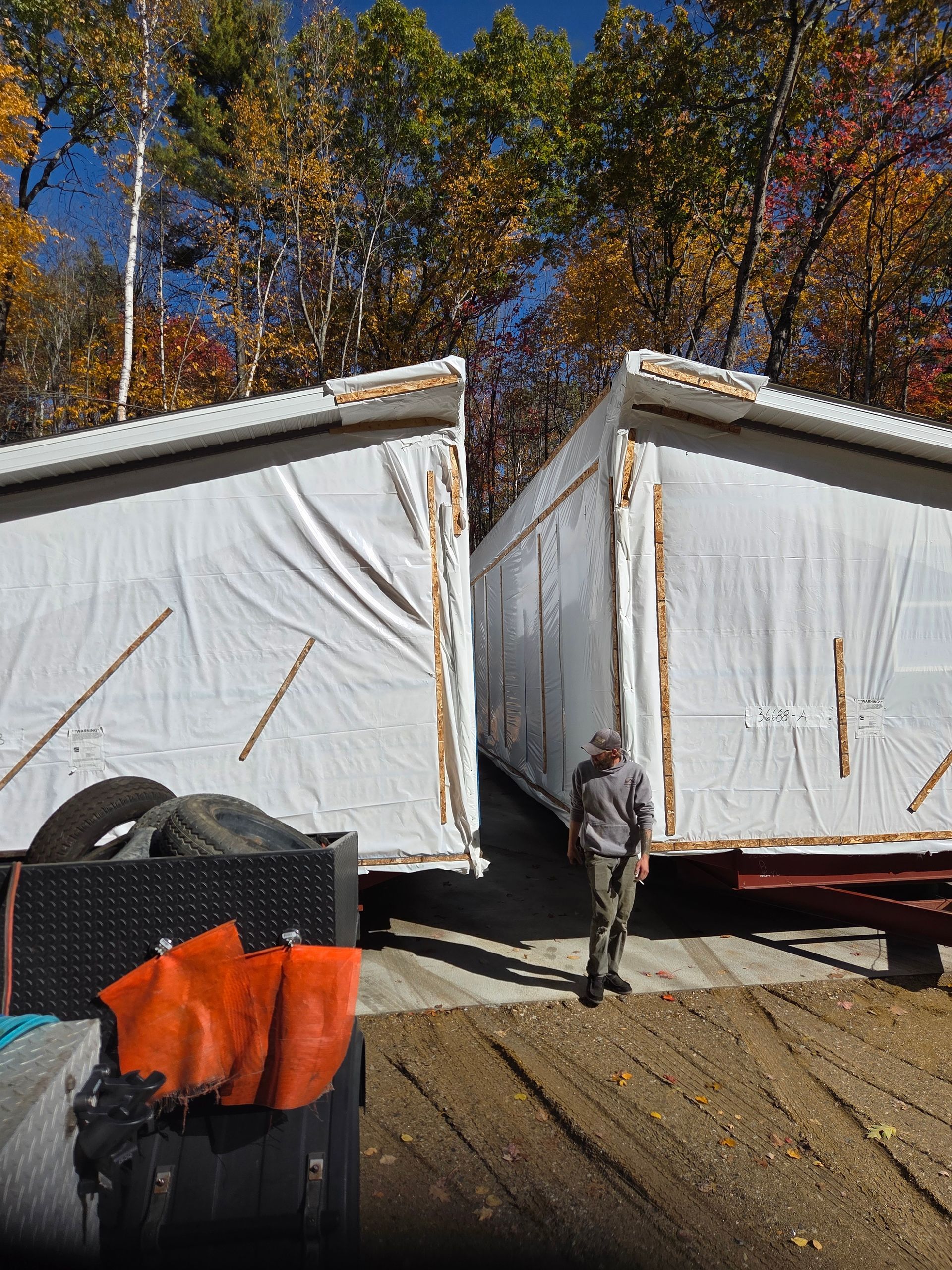 A person stands between two large, white-wrapped modular building sections outdoors amidst autumn trees.
