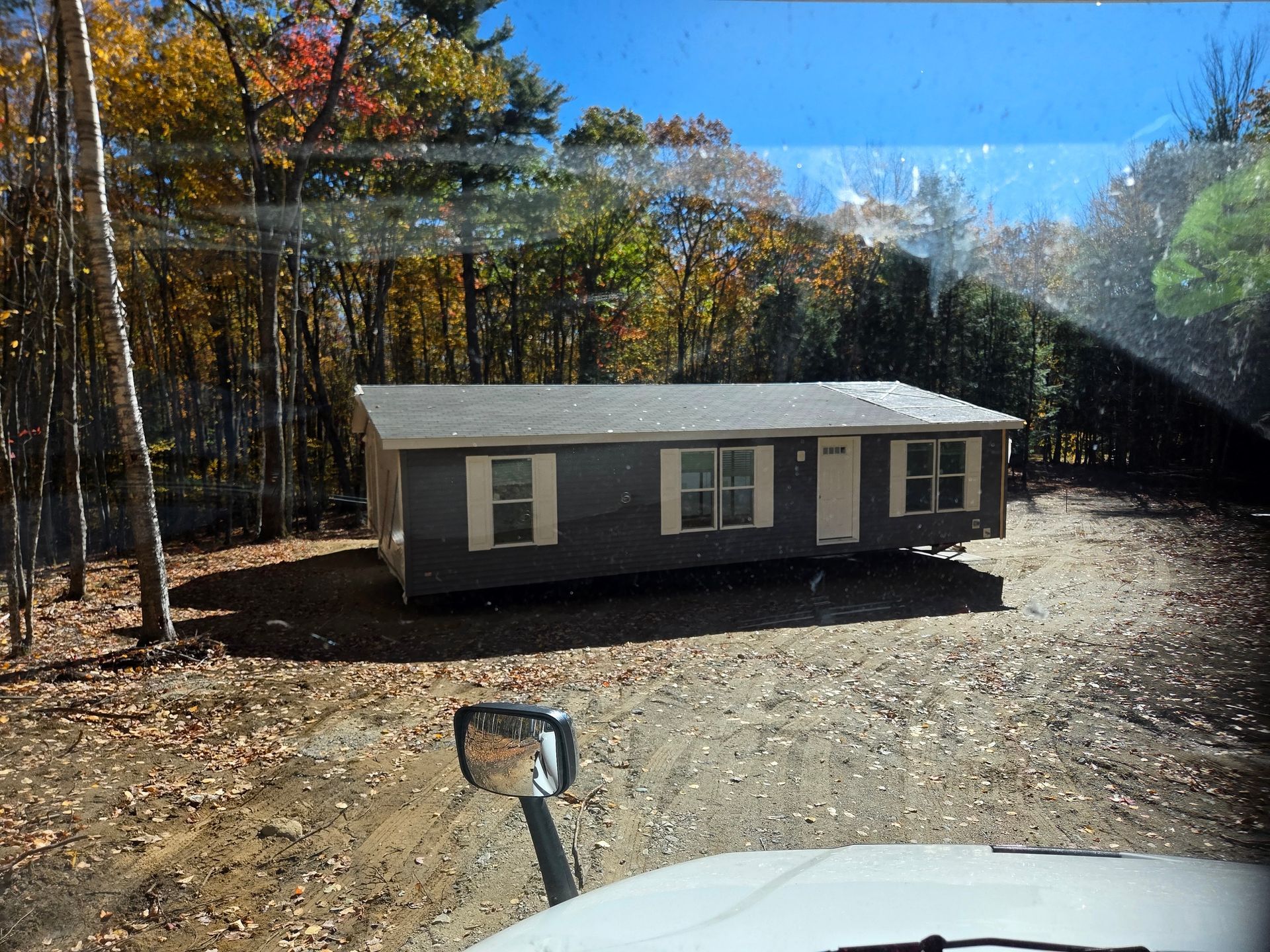 A grey manufactured home sits in a wooded, leaf-covered clearing, viewed from the cab of a vehicle.