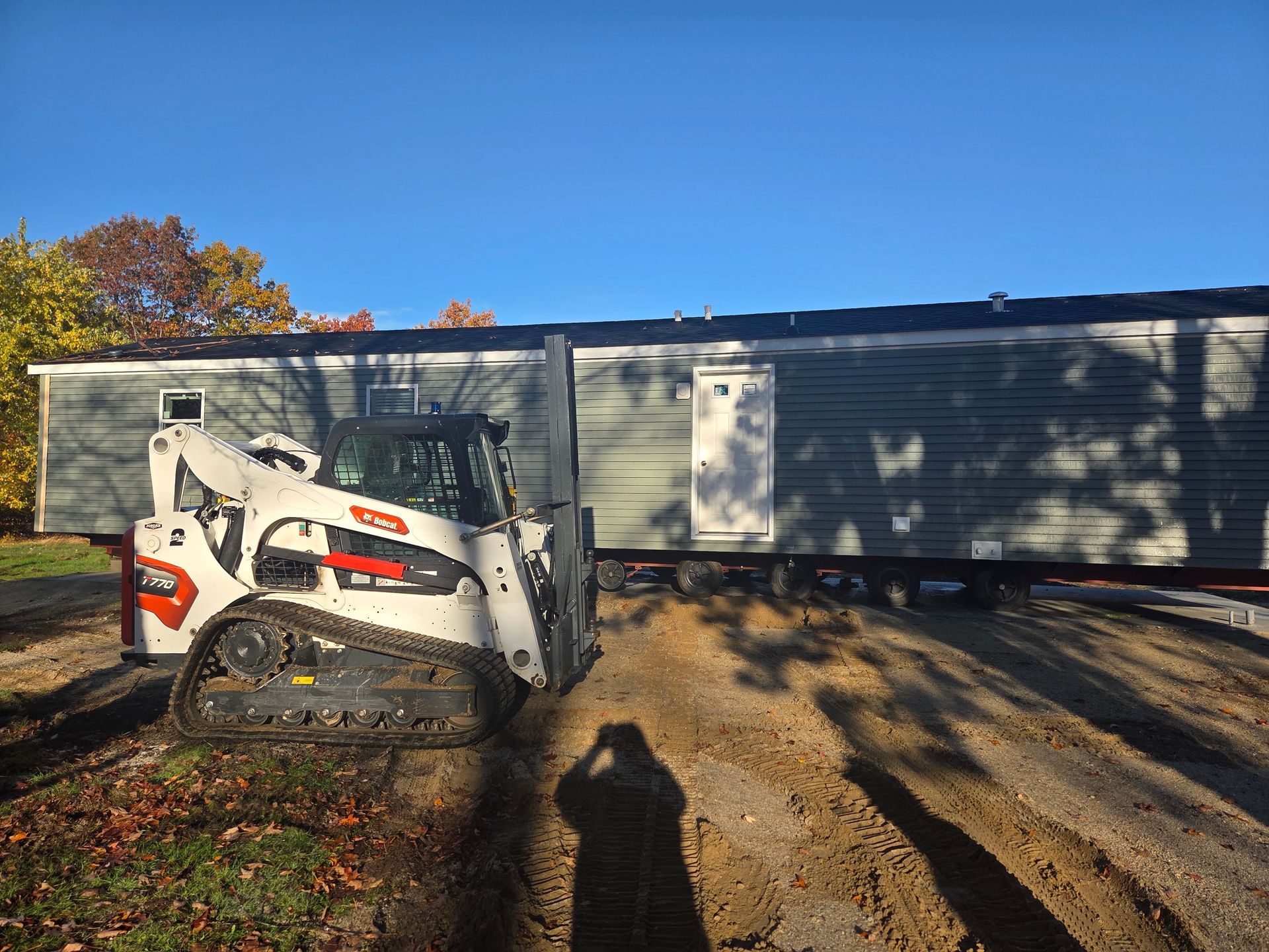 A white Bobcat compact track loader sits on a dirt site in front of a long, gray mobile home on a sunny day.
