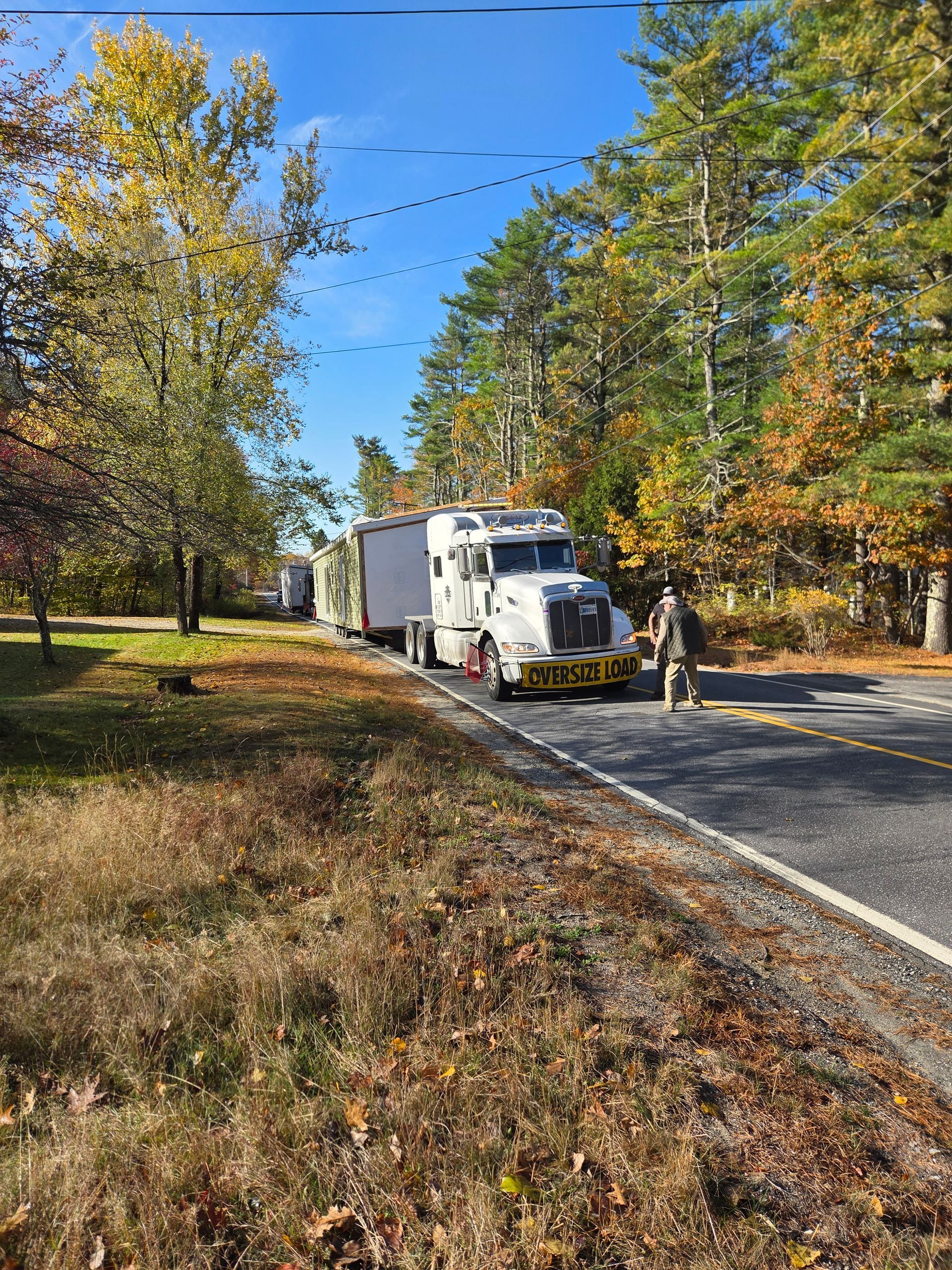 A white semi-truck with a large trailer parked on the shoulder of a tree-lined road with a person standing nearby.