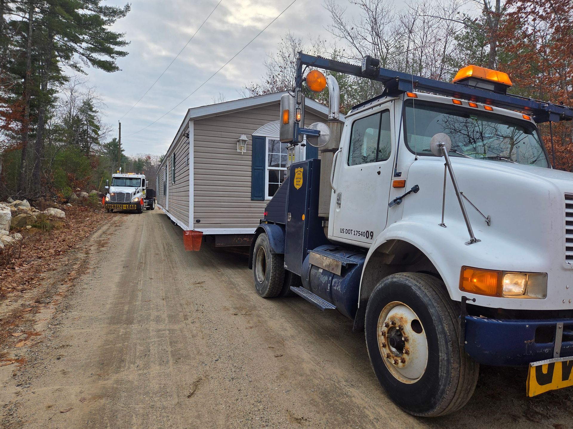 A large white semi-truck tows a tan mobile home down a gravel road lined with trees.