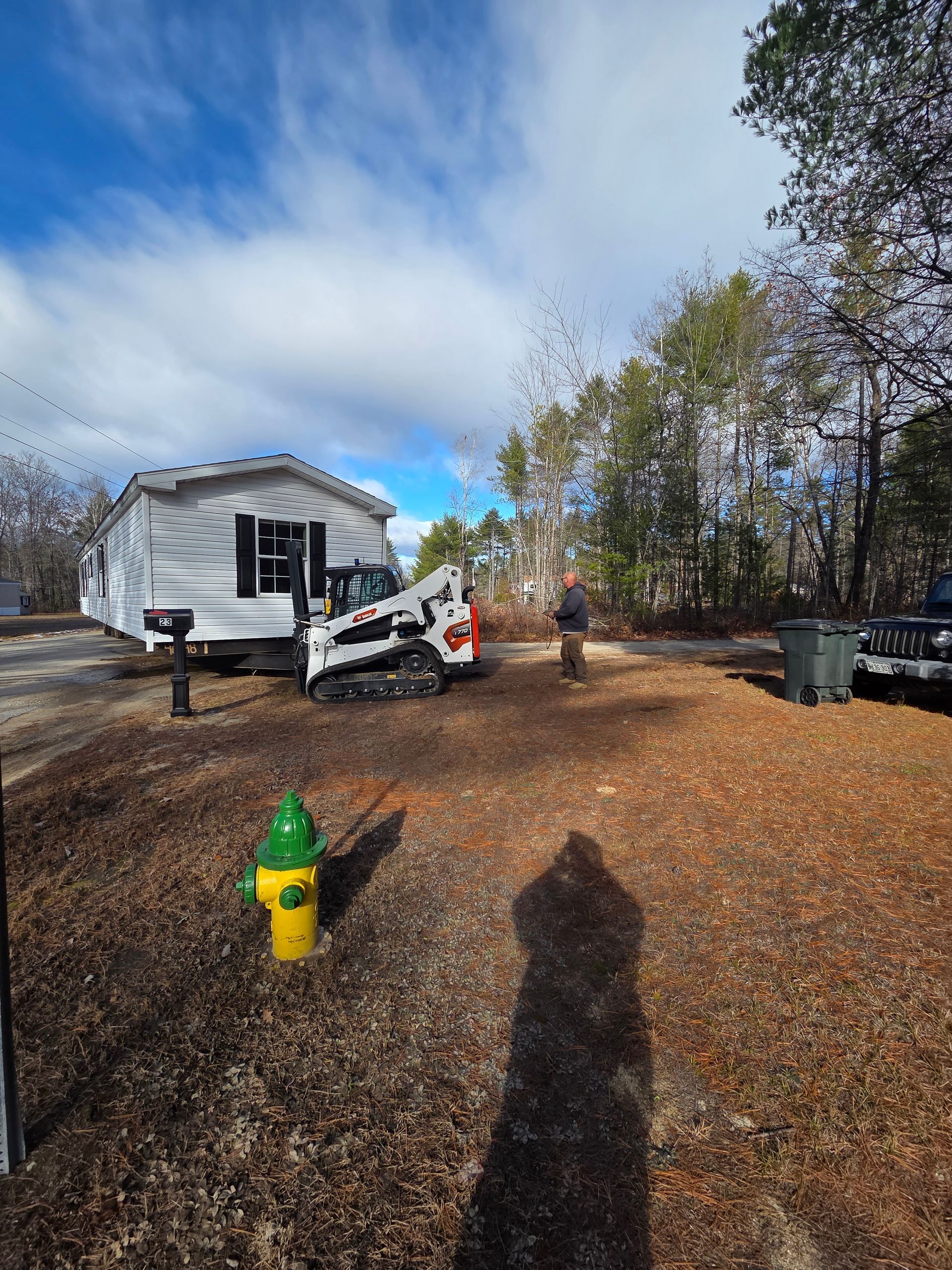 A white mobile home sits on a lot next to a skid-steer loader, a person, and a yellow fire hydrant on a leaf-covered yard.
