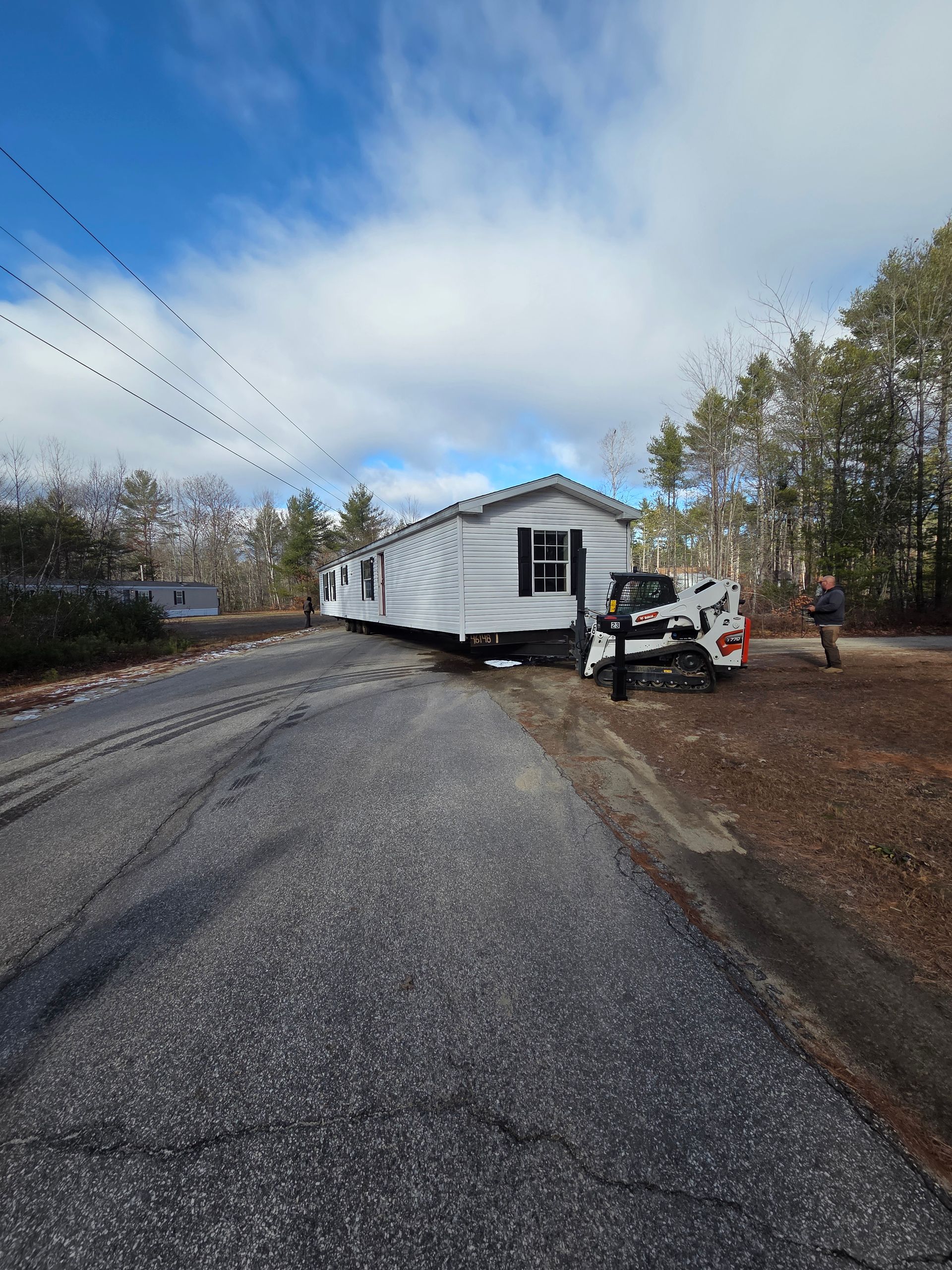 A white mobile home being transported along a rural road, pulled by a compact track loader.