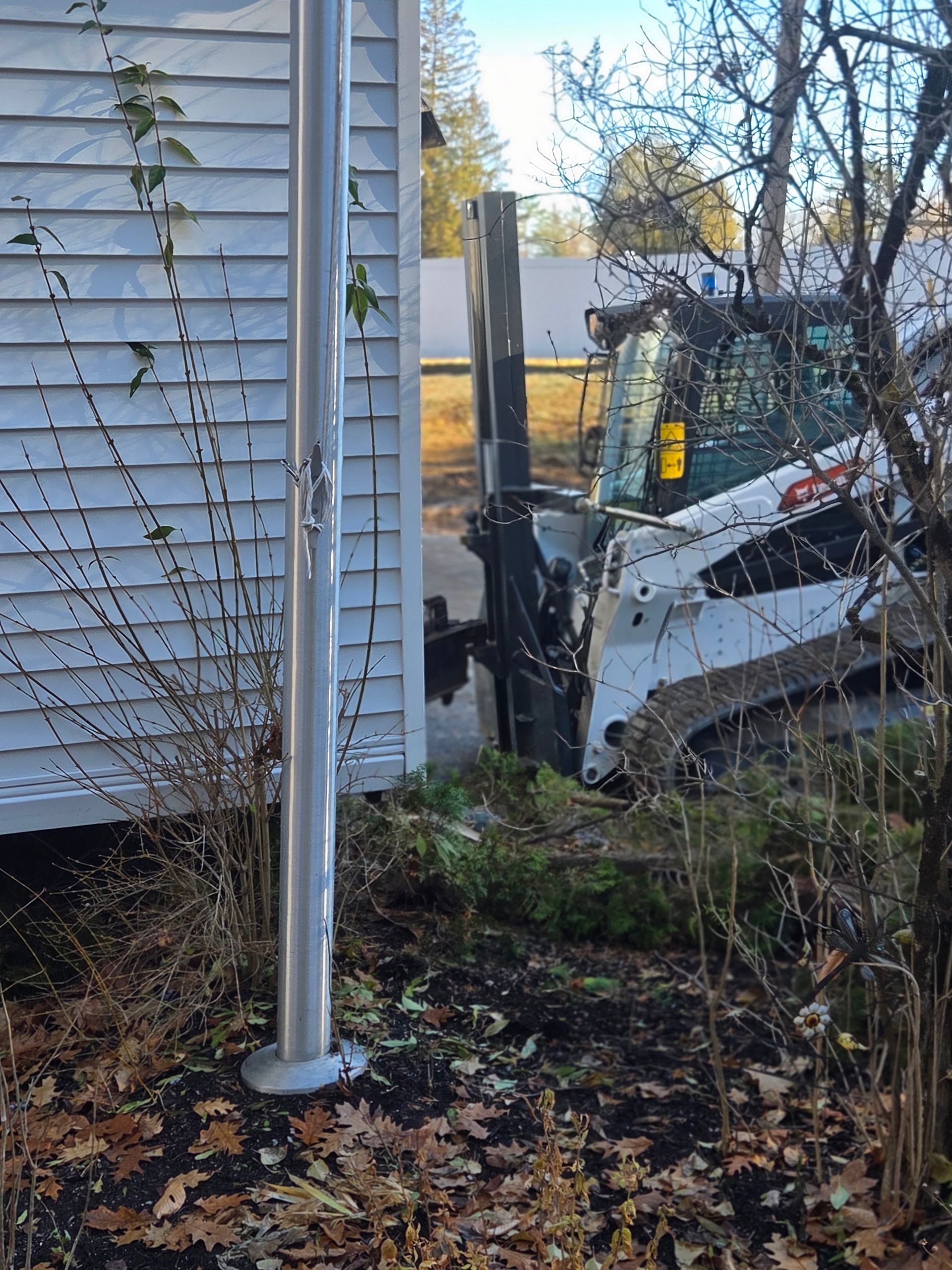 A white Bobcat skid-steer loader parked next to the corner of a white building with siding, partially obscured by bushes.