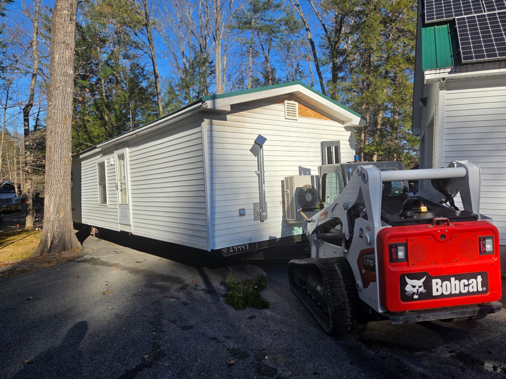 A white mobile home sits in a wooded area next to a red Bobcat skid steer loader on a gravel driveway.