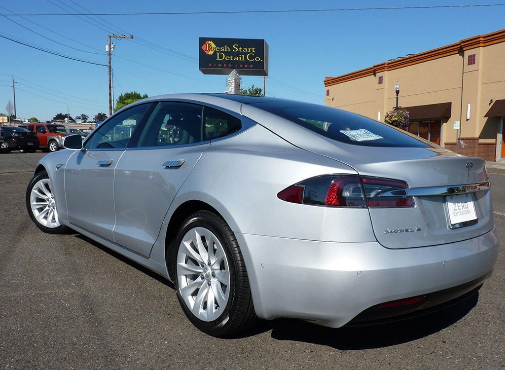 Silver Tesla sedan parked on a paved lot in front of a business on a sunny day.
