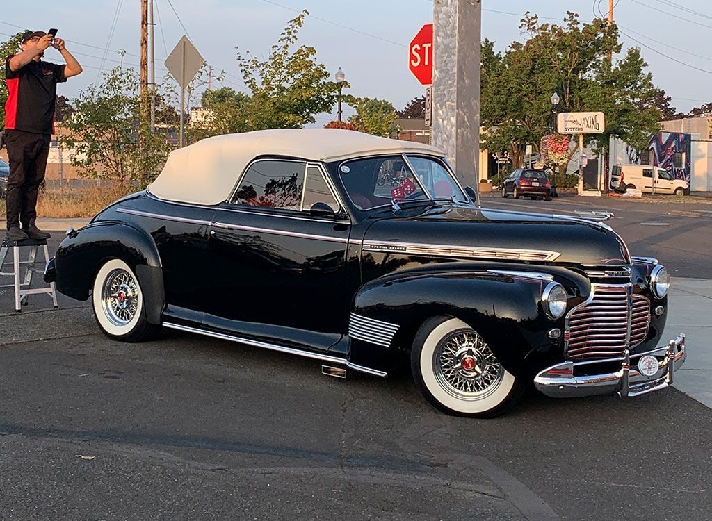 Black classic car, white top, parked near a stop sign. Photographer standing on a stool.