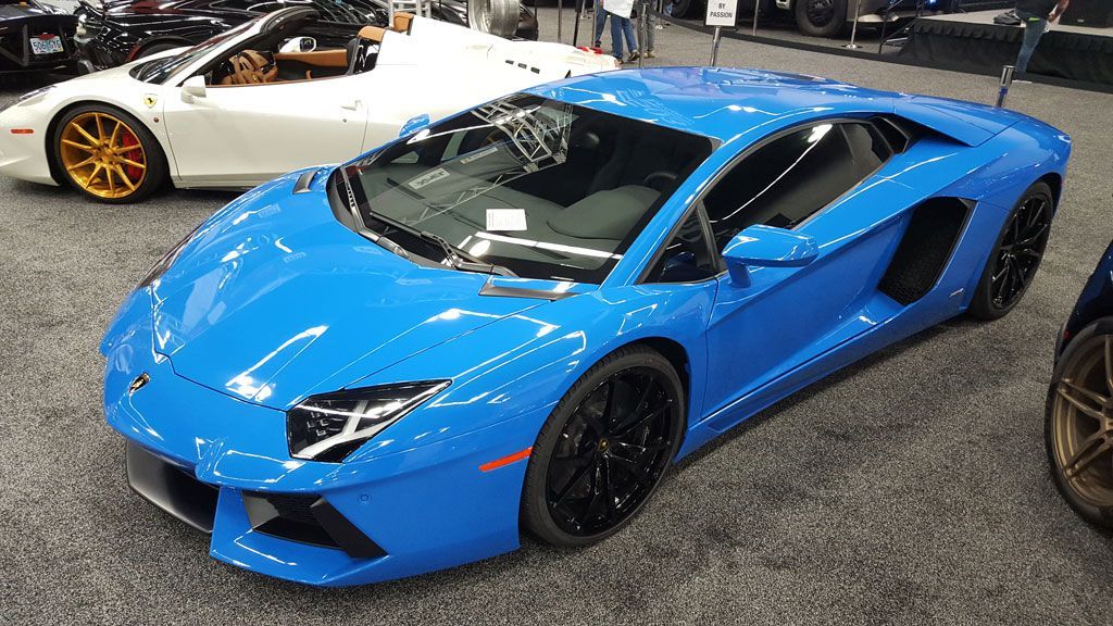 Blue Lamborghini Aventador sports car at a car show, black wheels, white Ferrari in background.