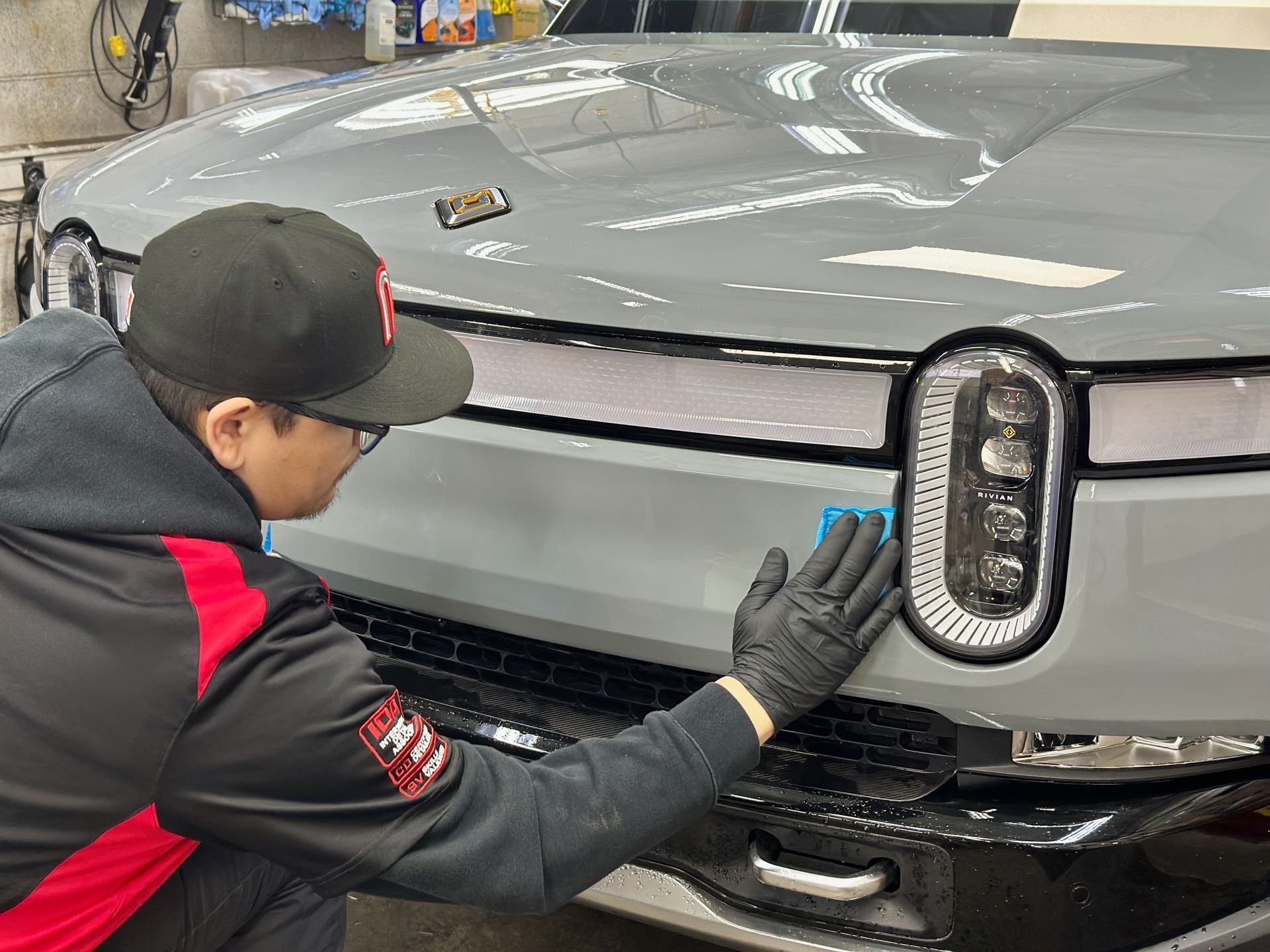 Man in black gloves applies a product to a gray electric truck's front bumper.