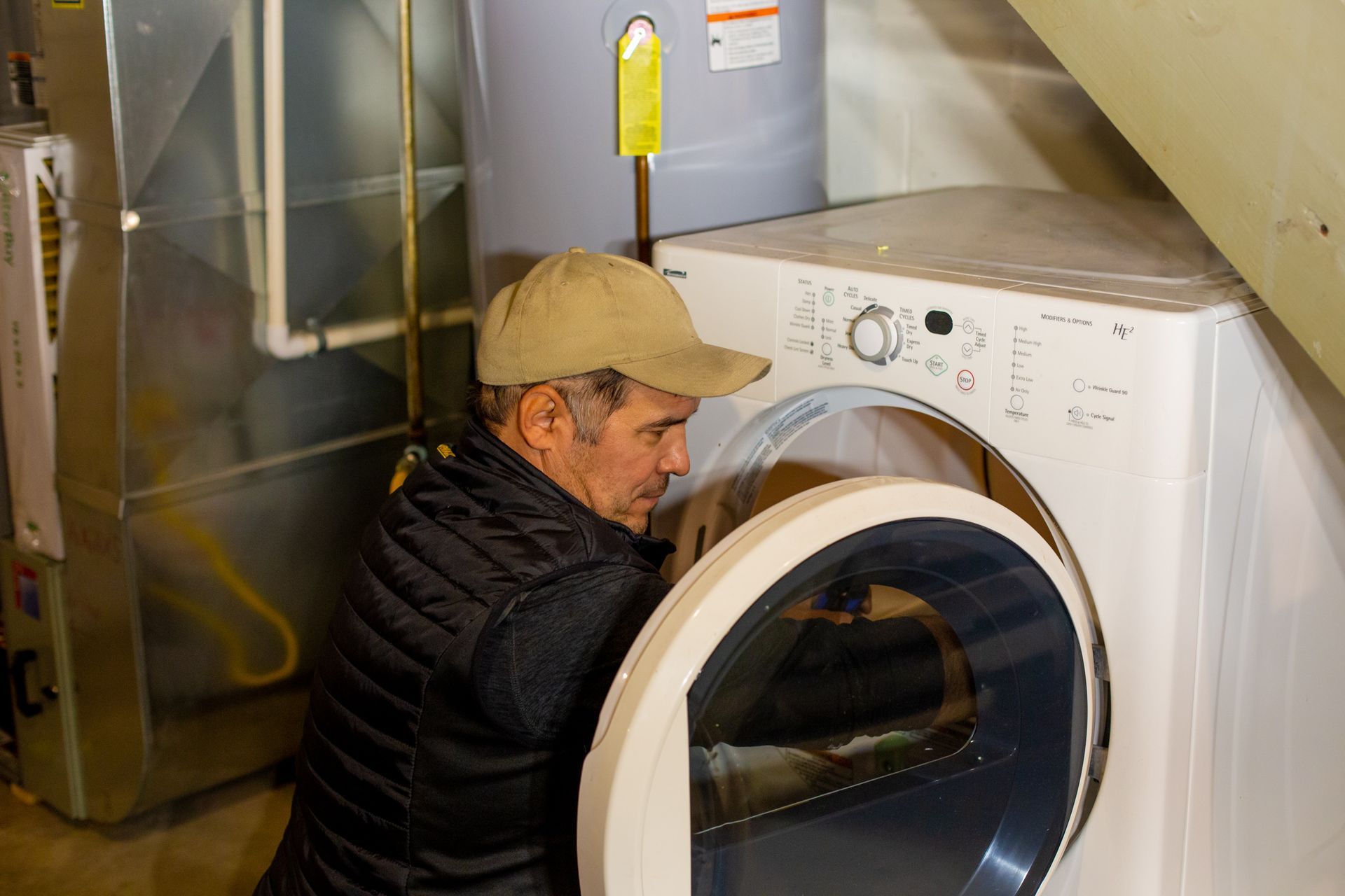 Man in cap leans into open dryer door in a basement, checking its interior.