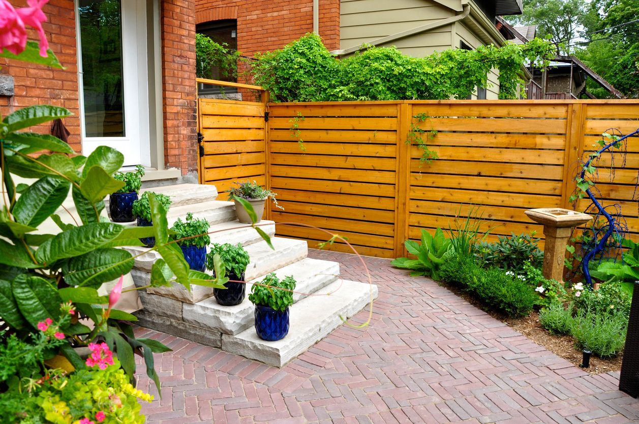 Brick garden path beside steps, potted plants, and a wooden fence in a lush backyard