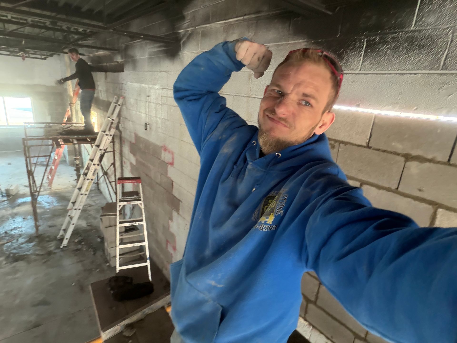 Person in blue hoodie taking a selfie in an unfinished room with ladders and construction materials.