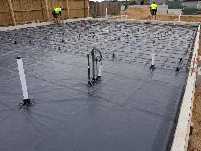 Two workers besides a foundation covered in poly-builder's film on a construction site.