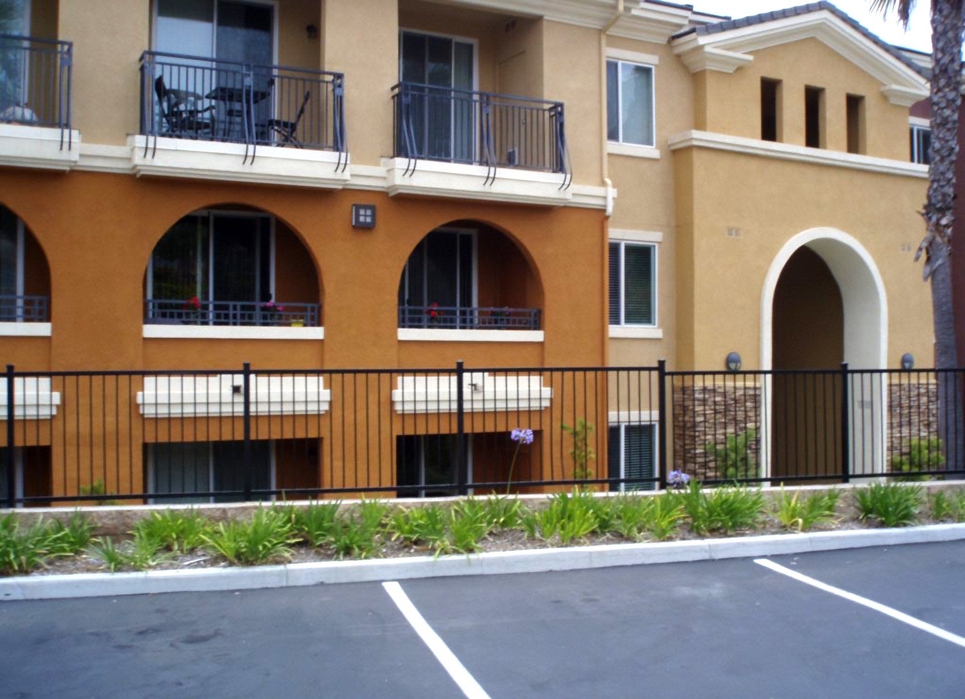 Tan stucco apartment building with arched balconies and a black fence beside a parking lot