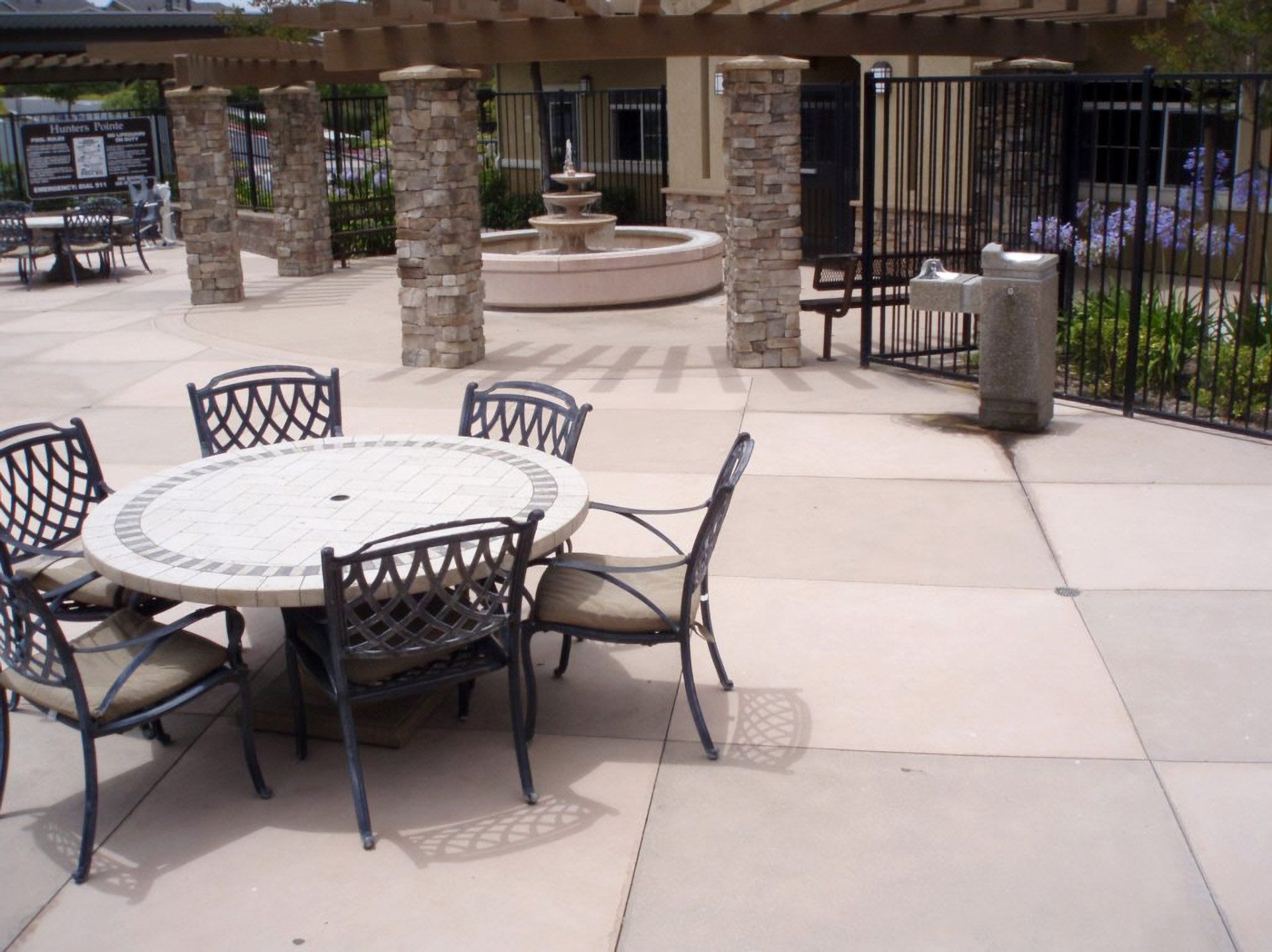 Outdoor stained patio with round table and chairs near a fountain and black metal fence