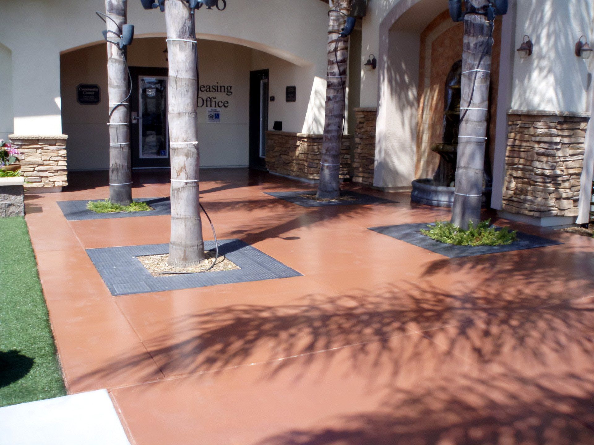 Sunlit courtyard with cororized concrete  walkway, palm trees, and stone arches beside a building entrance