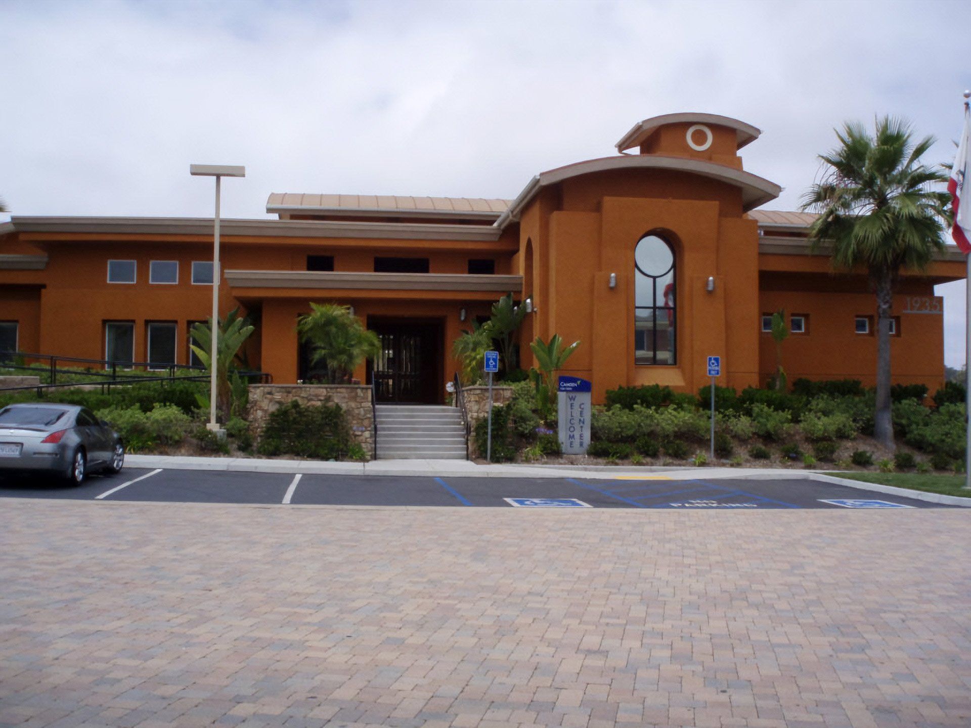 Orange stucco building with arched entrance, steps, stamped concrete, palm trees, and parking spaces in front.