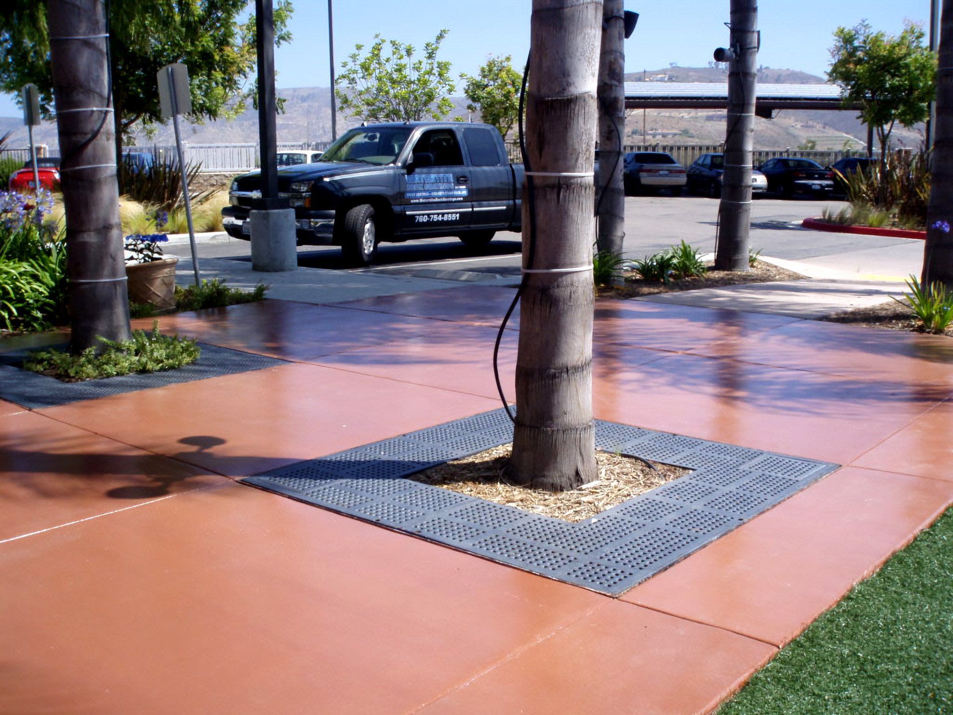 Shaded sidewalk with trees, orange stained waterproof pavement, a parked pickup truck, and a grassy edge in the background