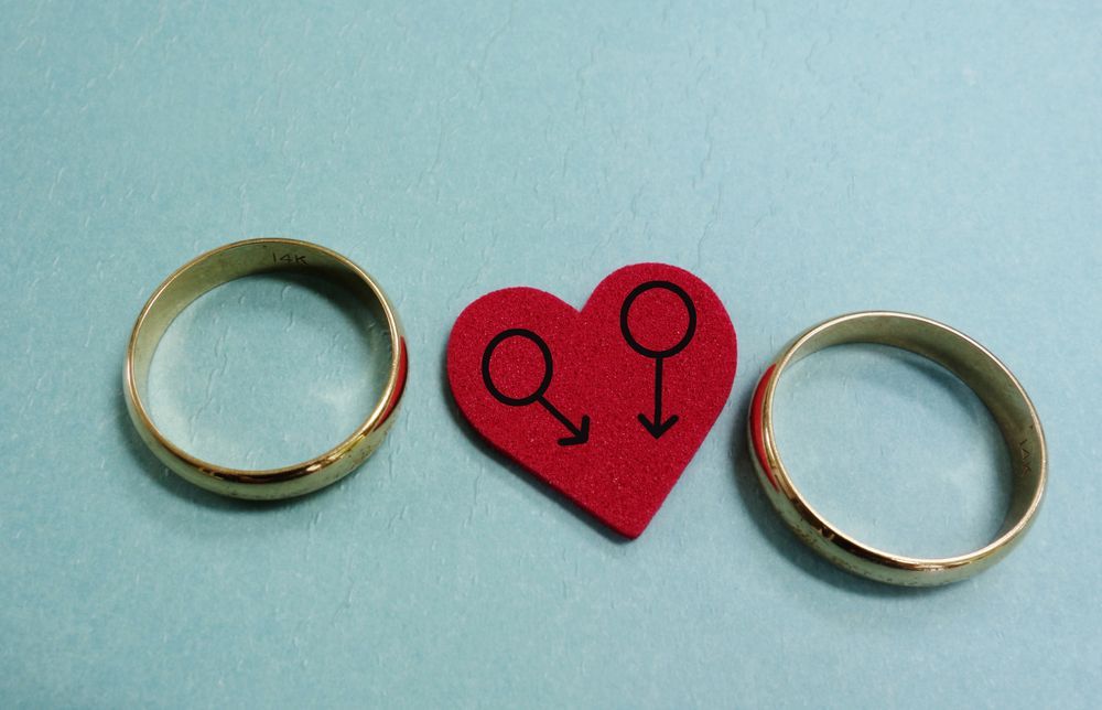 Two Gold Wedding Rings Flank a Red Heart With Two Male Symbols — Trapski Family Law Geelong in Newtown, VIC