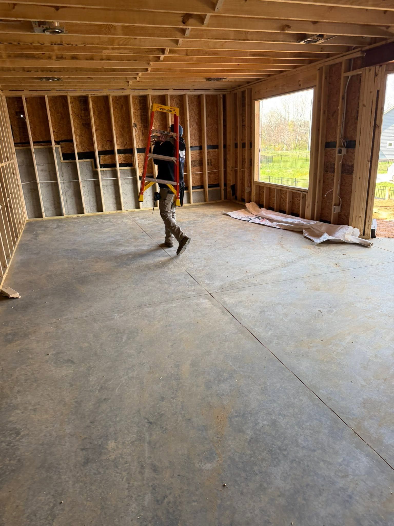 Worker inside unfinished wooden room with exposed studs and concrete floor, carrying tools.