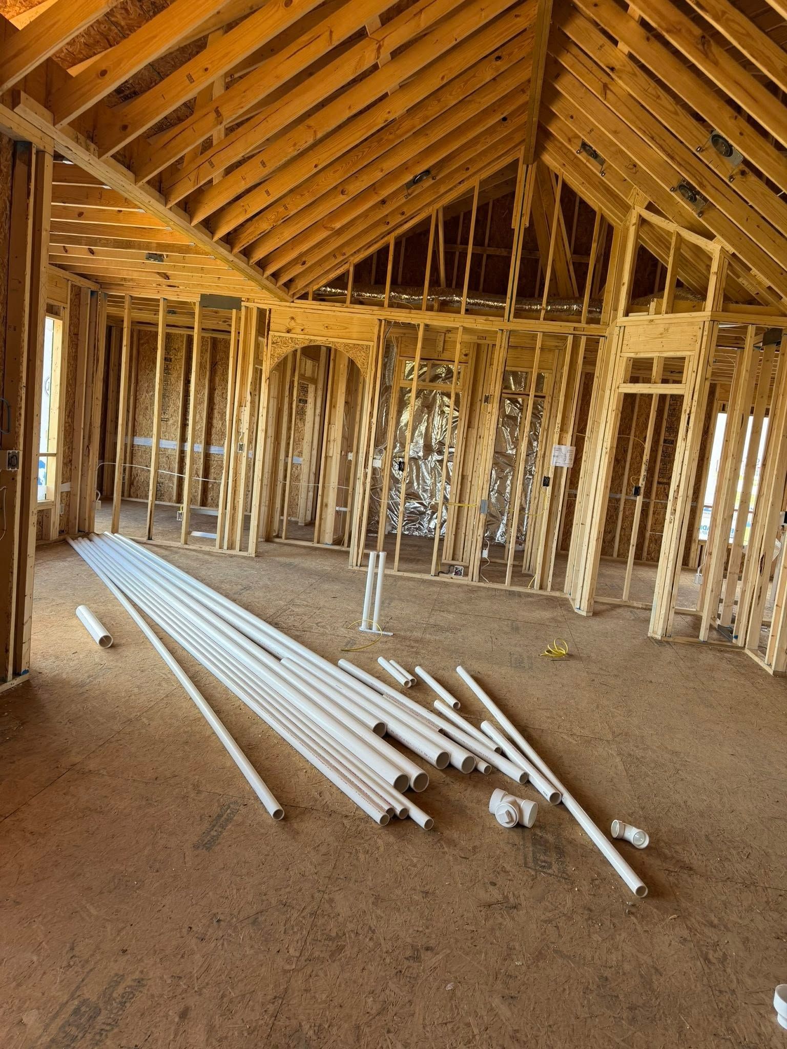 Unfinished house interior with exposed wooden framing and white PVC pipes on the dirt floor.