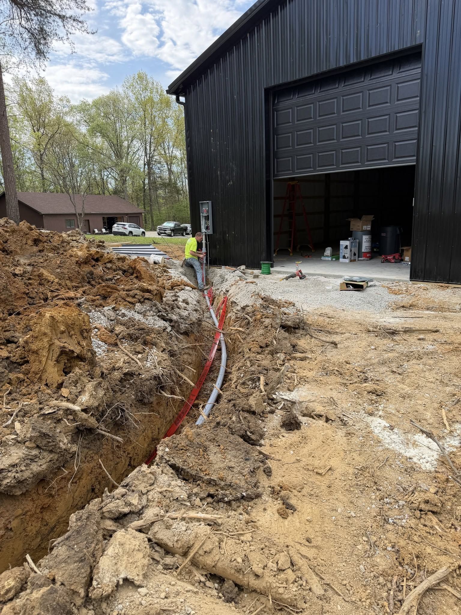 Excavation trench beside a black metal building, with muddy ground and an open garage door.