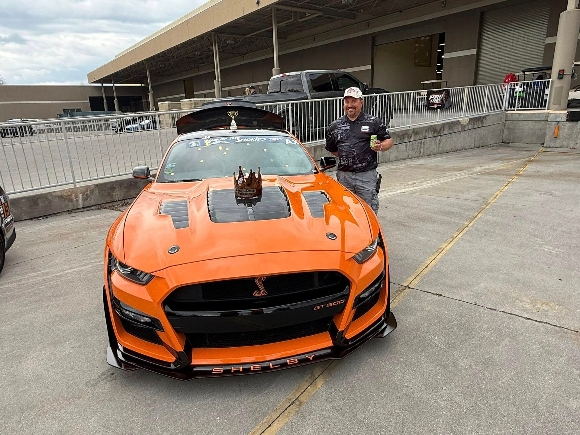 Orange sports car with large rear wing in a racetrack pit area, with a person standing beside it