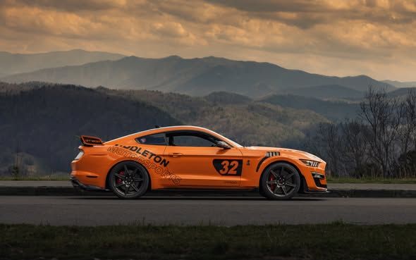 Orange race car with number 32 driving on a road, with mountains in the background.