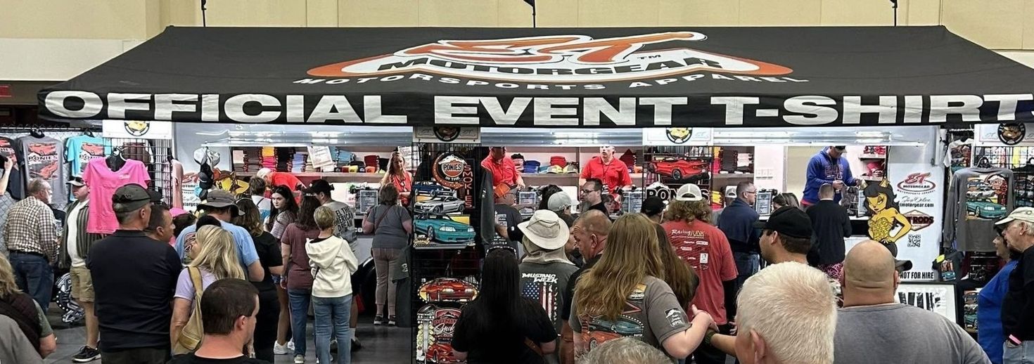 Crowded merchandise booth with a large “OFFICIAL EVENT T-SHIRT” sign and people browsing shirts and souvenirs
